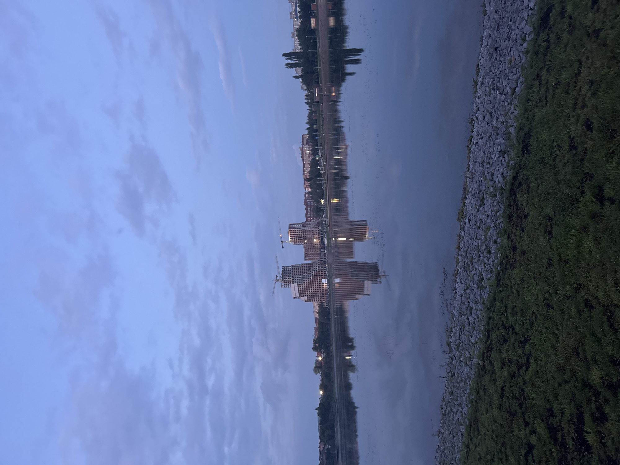 This is a photograph of a dusk scene at the Ufa waterfront, located in Ufa, Russia. In the middle ground, a body of water reflects the twilight sky and the city skyline, which features several tall buildings under construction, some with cranes visible. Along the waterfront, there is a park area with trees and lampposts. The foreground shows a grassy embankment leading down to a rocky shore. The sky is a dusky blue with scattered clouds.