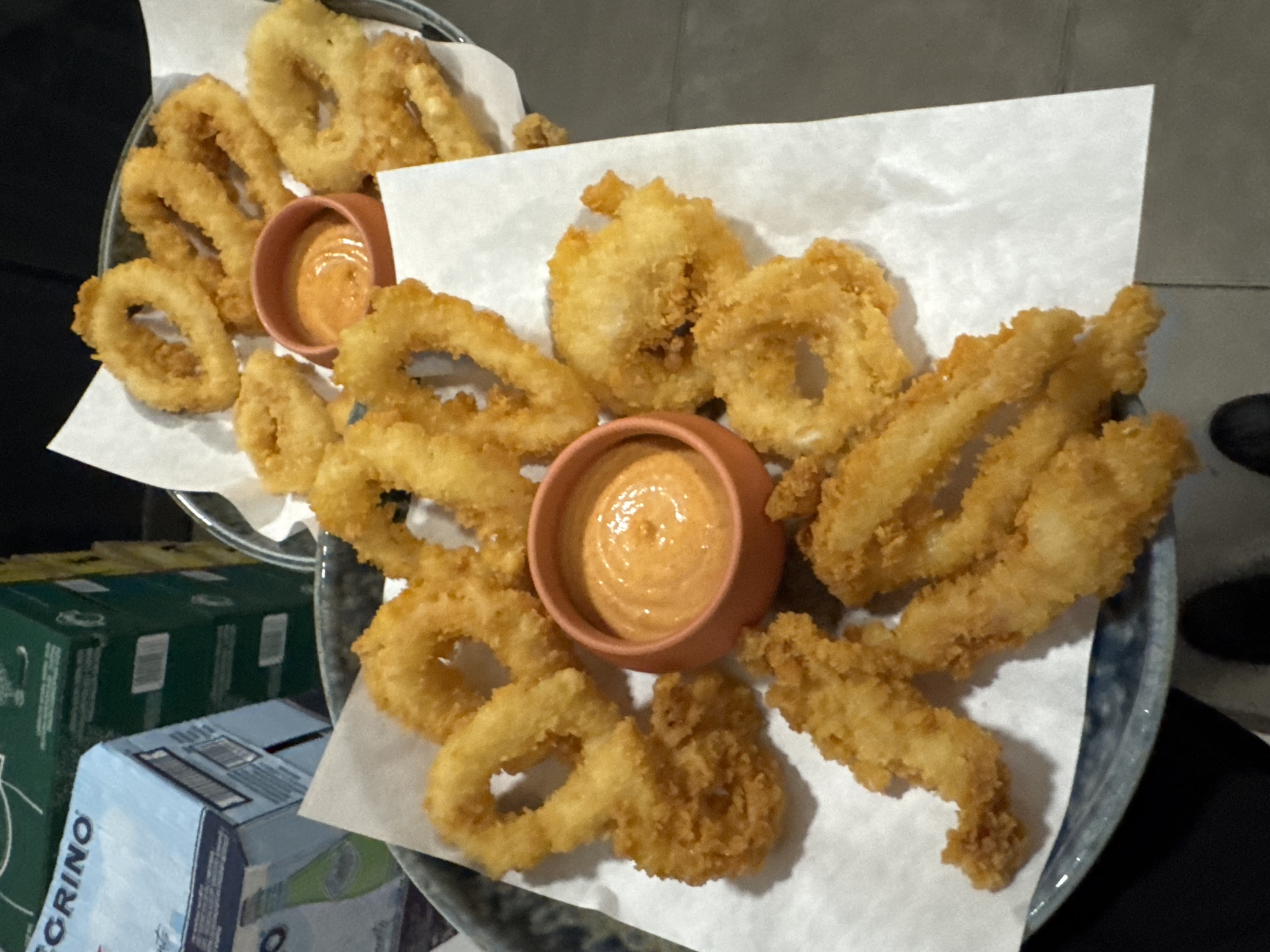 The image displays a close-up, eye-level shot of a serving of golden-brown fried calamari, presented on white parchment paper within a dark-rimmed plate or shallow bowl. The platter contains numerous rings and irregular pieces of fried seafood, characterized by a crispy, textured coating. Two small, terracotta-colored ceramic bowls are embedded among the food, each holding a dollop of creamy, orange-hued dipping sauce with a slightly swirled surface.
In the blurred background, stacked cardboard boxes are partially visible. A prominent light-blue box on the lower left features the white text "S.PELLEGRINO," indicating a well-known sparkling water brand. Another dark green box is visible behind it. The lighting is subdued and even, suggesting an indoor setting like a restaurant, bar, or home dining area. No people are visible, and the focus is entirely on the food presentation, ready for consumption. The scene suggests a casual dining experience in Sofia, Bulgaria.