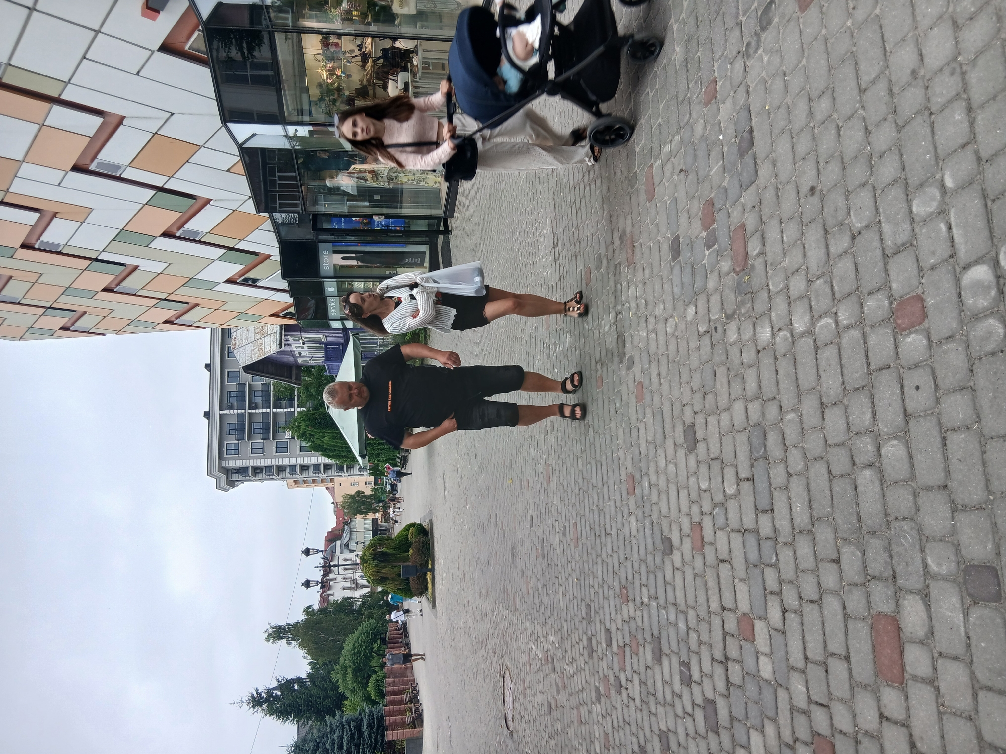 In Truskavets, Ukraine, a cloudy day unfolds on a cobblestone street. A man in a black t-shirt and shorts stands with his hands on his hips, looking to his left. Beside him, a woman with dark hair, wearing a striped shirt and black skirt, walks away from the camera, carrying a white bag. Further down the street, another woman in a light pink sweater pushes a stroller with a baby inside. The background features modern buildings with geometric facades and glimpses of shops. Green trees and bushes line the street, suggesting a well-maintained urban park or pedestrian area. The overall atmosphere appears to be a casual stroll on a day with overcast skies. There is no visible text in the image.