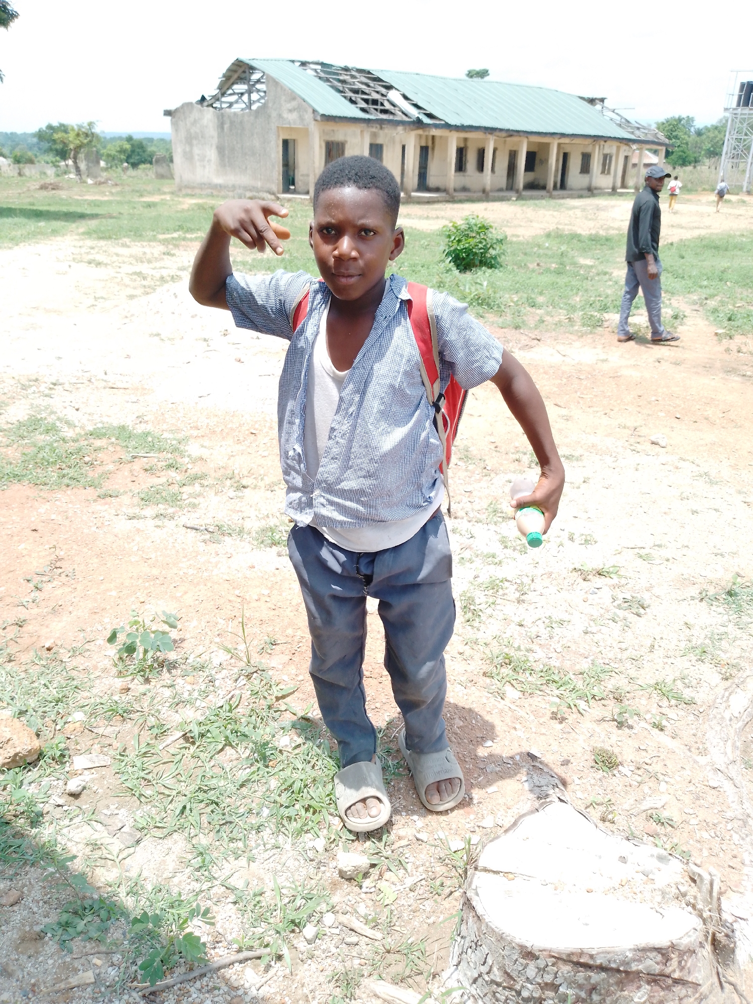 A young boy stands in a dry, grassy field in Akwanga, Nigeria. He wears a blue striped shirt over a white undershirt, dark pants, and light-colored sandals. A red backpack is on his back. In his right hand, he holds a small bottle. His left hand is raised in a gesture. Behind him, a partially dilapidated, one-story building with a green roof and missing sections is visible. Another man walks in the background. A cut tree stump is in the foreground.