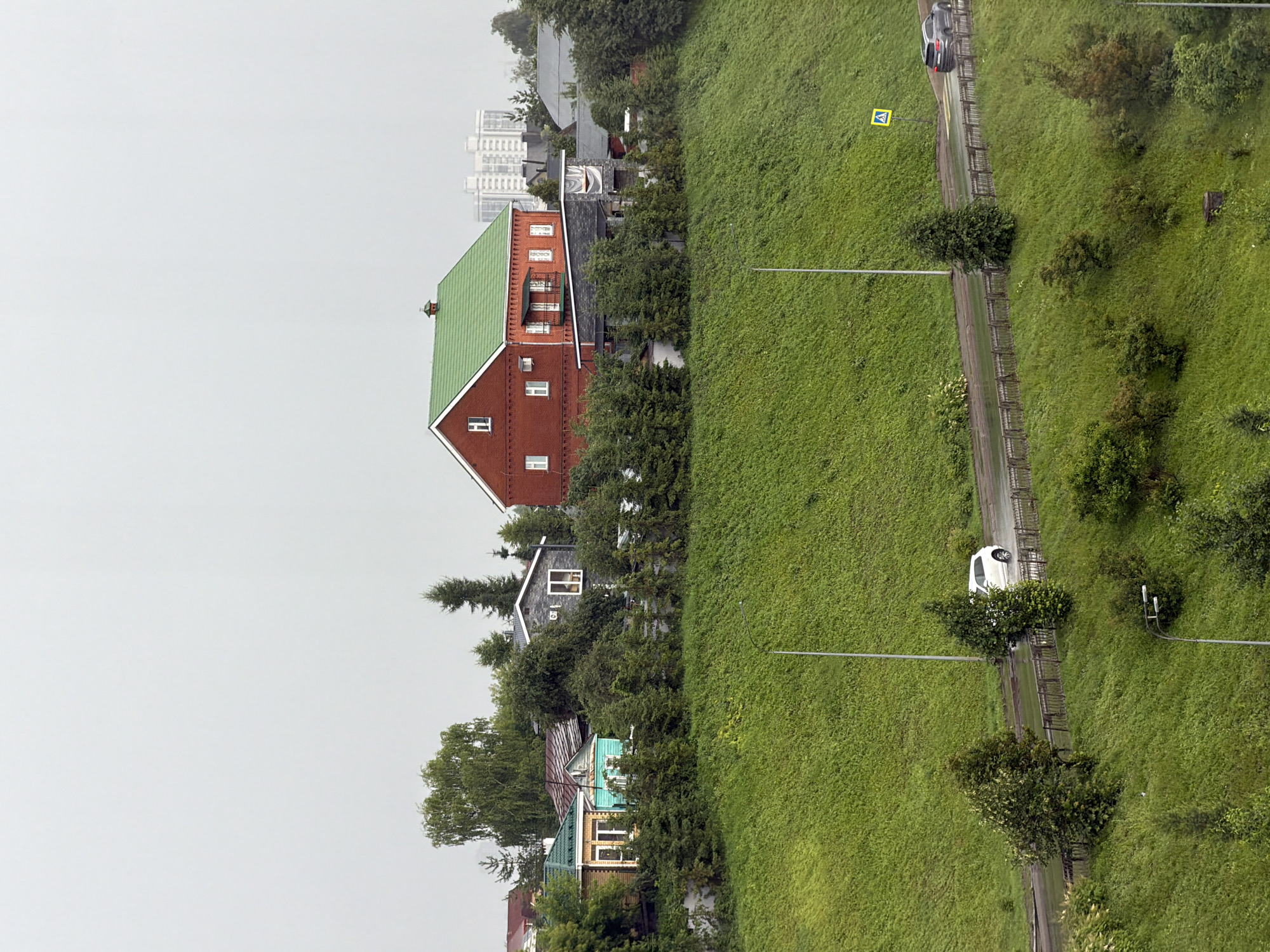 The image depicts a hillside residential area in Kazan, Russia, under overcast skies. A multi-story red brick house with a green roof is prominently featured, surrounded by trees. Below the hill, a wet asphalt road runs horizontally, with two cars visible. A white car is driving away from the viewer on the left side of the road, while a dark-colored SUV is driving towards the viewer on the right. A pedestrian crossing sign is visible on the right side of the road. The hillside is covered in lush green grass and scattered trees. The overall impression is of a residential neighborhood on a damp, cloudy day.