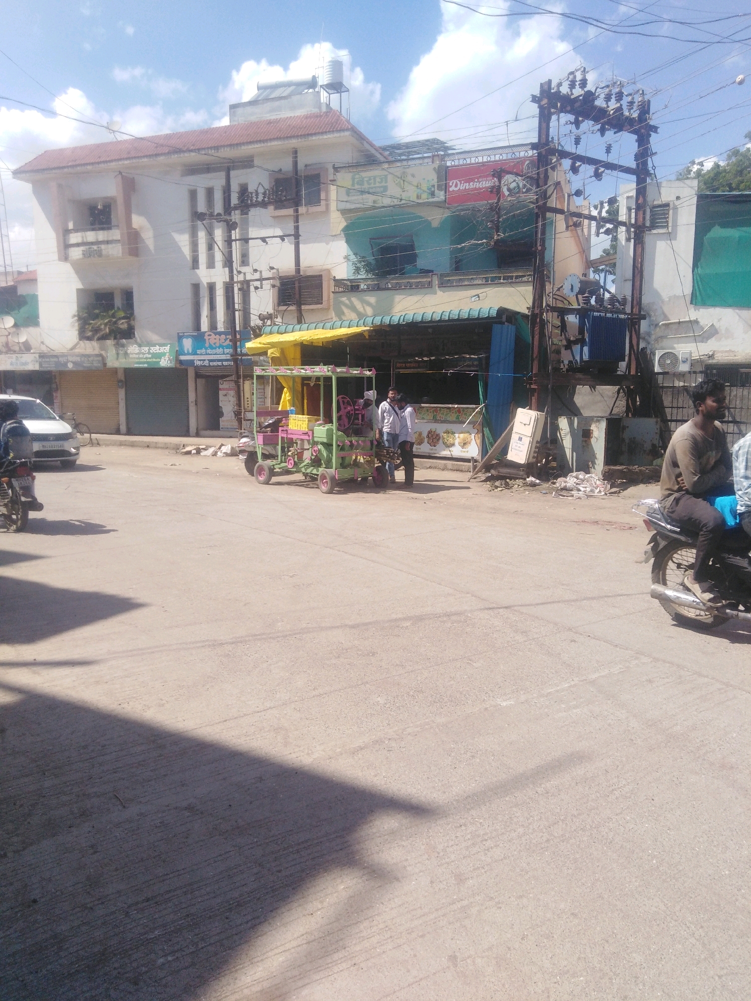 The image captures a street scene in Nanded, India, under a partly cloudy sky. In the foreground, the asphalt road shows tire tracks and shadows from overhead structures. A white car is parked on the left side of the road.
Dominating the midground is a pink and green cart with a generator and what appears to be a juice-making machine, manned by two individuals in light-colored shirts. The cart is positioned in front of a building with a green awning, beneath an electrical transformer and utility pole.
Further back, a multi-story building with red-tiled roofing features several windows and balconies. Various shopfronts with awnings line the street. Signs with Indian script are visible on some of these establishments, including one with "Dinsurha" written in red.
On the right side of the image, a man in a brown shirt is seated on a motorcycle, looking to the right. Another person in a checkered shirt is partially visible on the motorcycle behind him. The presence of electrical infrastructure, shops, and vehicles suggests a commercial or residential street in an urban setting. The overall impression is of a typical day in a busy Indian city.