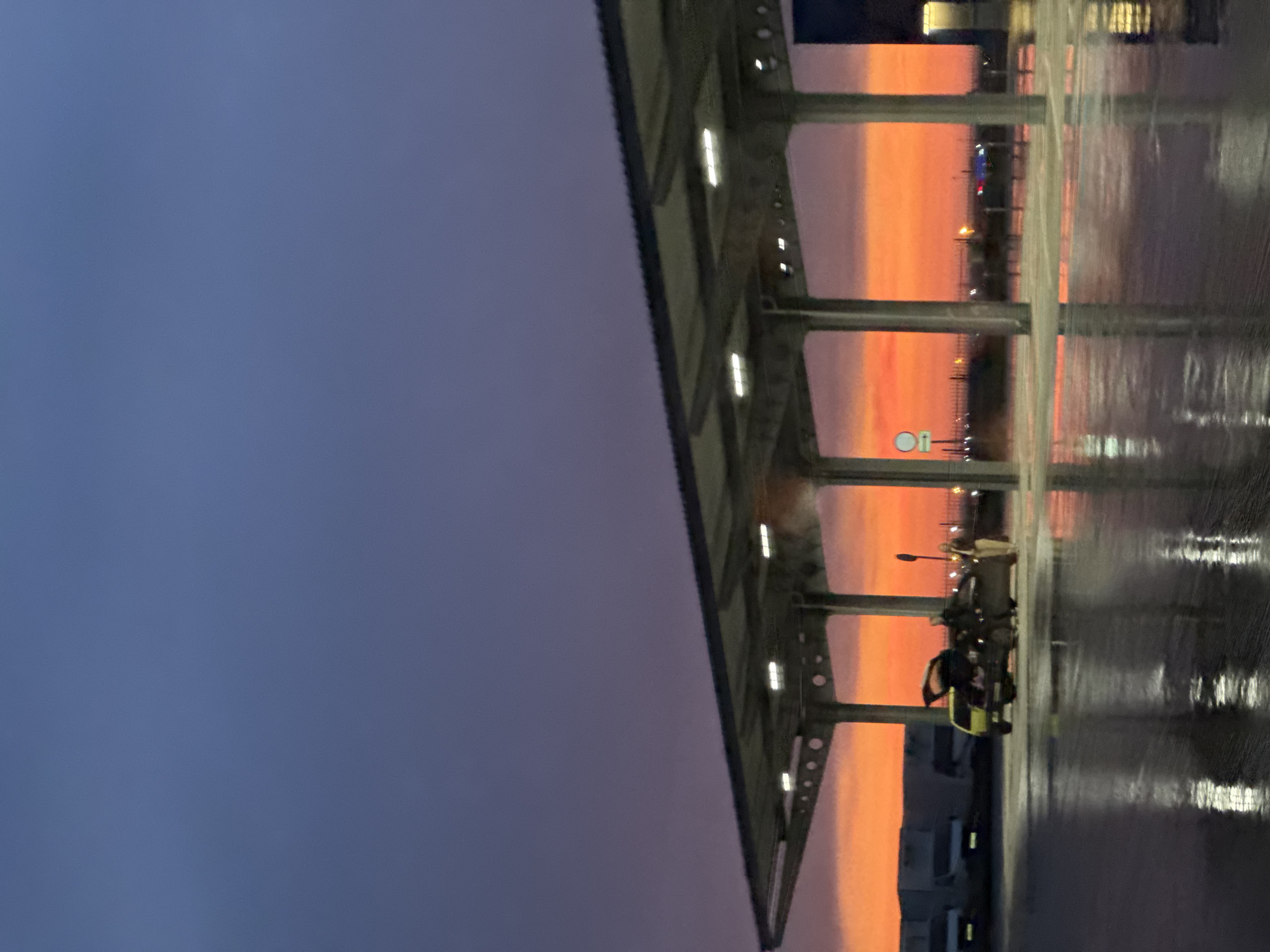 The image displays an outdoor scene at dusk or dawn, characterized by a dramatic sky featuring a deep indigo-purple upper region gradually transitioning to a vibrant orange-red at the horizon. The ground, likely asphalt or concrete, is entirely wet and highly reflective, mirroring the sky, structures, and illumination.
A large, modern canopy structure with a flat roof and multiple support pillars extends across the mid-ground. Several white rectangular light fixtures are active on the underside of the canopy. Beneath this structure, in the center-left, two vehicles are present. On the left is a light-colored hatchback or small SUV with its rear hatch open. Adjacent to it, on the right, is a darker-colored sedan. Two individuals are visible near the darker sedan: one person in dark clothing stands near the rear of the vehicle, while another person in lighter clothing stands further to the right. A white circular sign, possibly regulatory, is mounted on a pole behind the vehicles.
In the background, to the left, several large, dark, industrial or commercial buildings are visible, with multiple white vans parked near them. A section of another building is partially visible on the far right. Fencing and distant lights are discernible along the horizon. The wet ground and active artificial lighting suggest recent precipitation and low natural light conditions. The location is in the city of Unknown, Unknown.