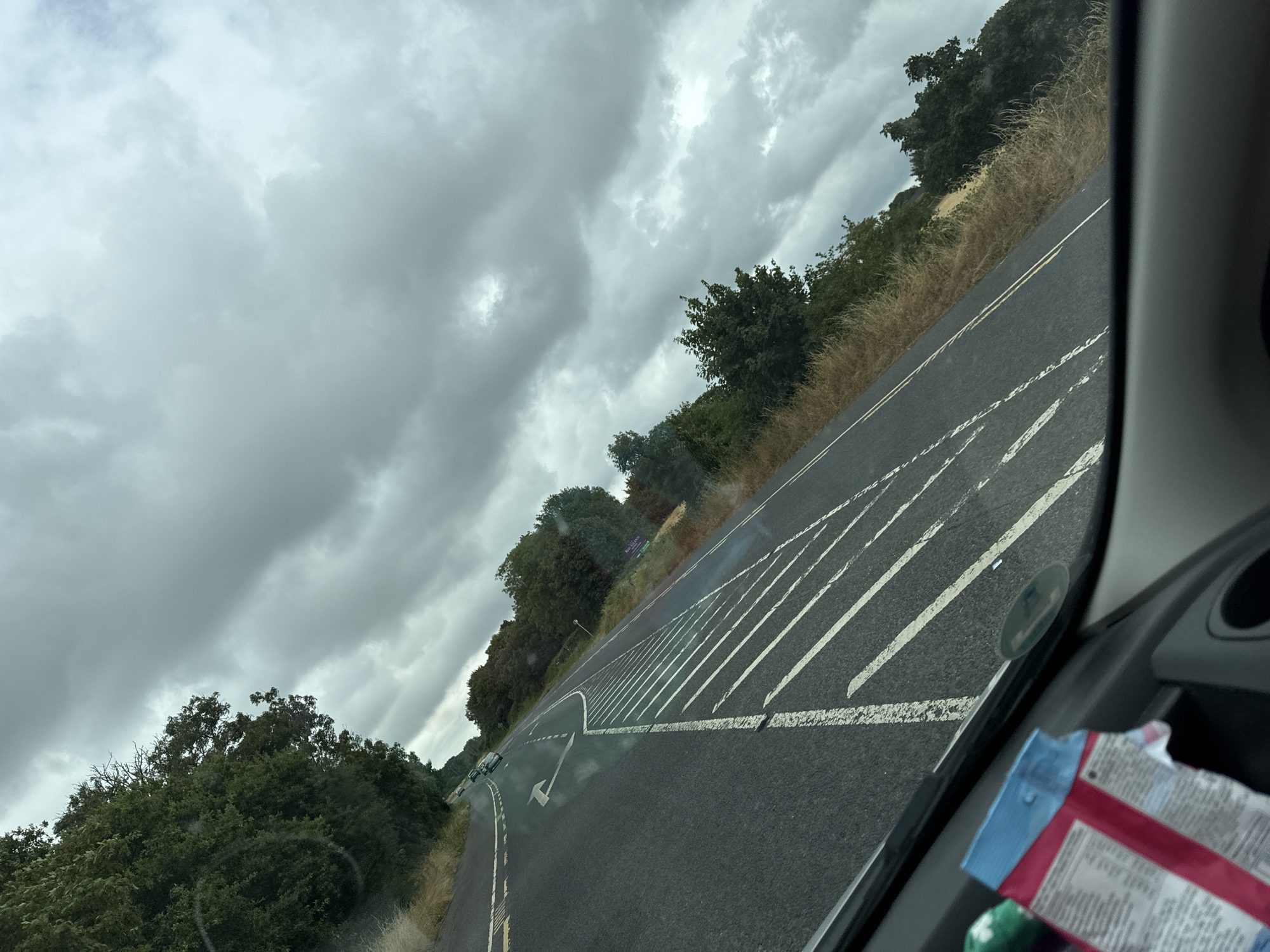 The image shows a view from inside a vehicle, looking out at a two-lane road under a cloudy sky. The road surface is asphalt and marked with white lines. On the right side of the road, there are white markings that appear to be directional arrows and hatching. The left side of the road is bordered by dry grass and trees. In the distance, on the left side of the road, there are a few vehicles on the road. The sky is filled with grey and white clouds, suggesting overcast weather. The overall scene depicts a rural or semi-rural road environment.