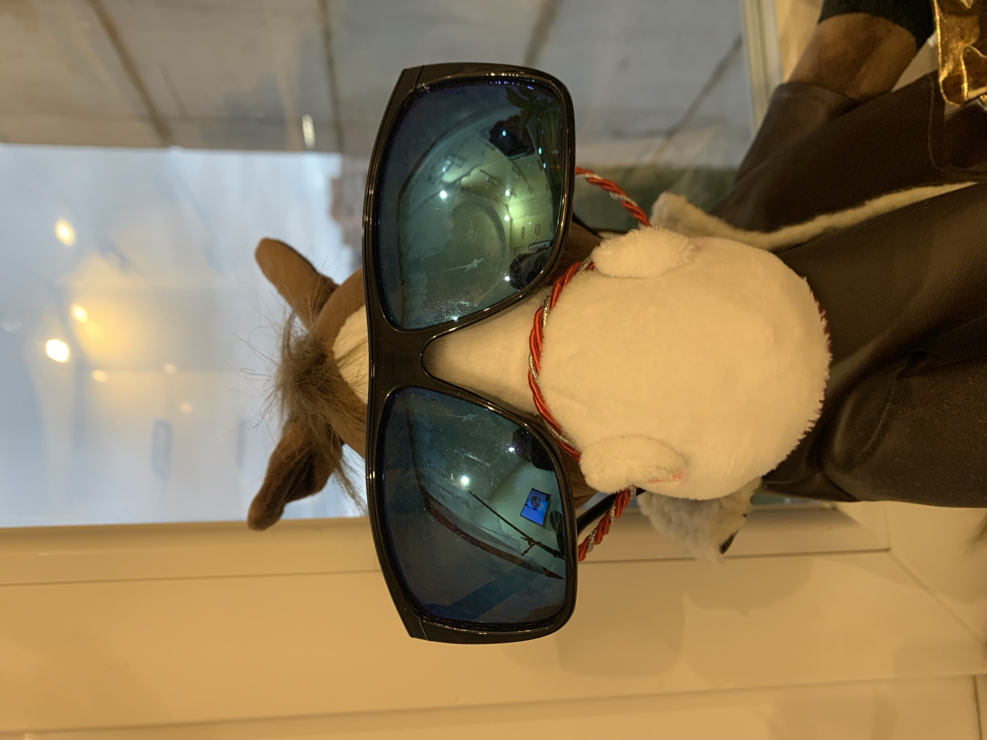 An indoor, close-up photograph features a plush toy, likely resembling a reindeer, positioned in front of a window. The image, rotated to appear upright, prominently displays the toy wearing oversized, dark-framed sunglasses with highly reflective blue lenses. The reindeer-like toy has light brown furry ears and parts of its body, a white face or snout, and is adorned with a red and white braided string around its neck. It also appears to be wearing a dark brown garment, possibly a jacket or suit, with a hint of white trim.
The reflective blue lenses of the sunglasses offer a detailed glimpse into the immediate indoor environment where the photo was taken. In the right lens, reflections show multiple artificial lights and structural elements, along with a distinct small blue rectangular screen displaying white lines, suggestive of a computer monitor or digital display. The left lens reflection similarly captures indoor lighting and architectural features.
Through the window behind the toy, a blurred background is visible, dominated by light, neutral tones, indicating either an overcast day outside or reflections of interior lighting from another part of a building. The window frame itself is a light, neutral color. The setting appears to be an indoor space in Severodvinsk, Russia, where the toy is placed near a window, capturing detailed reflections of its surroundings. No specific activity is depicted, and no discernible text is visible within the image.