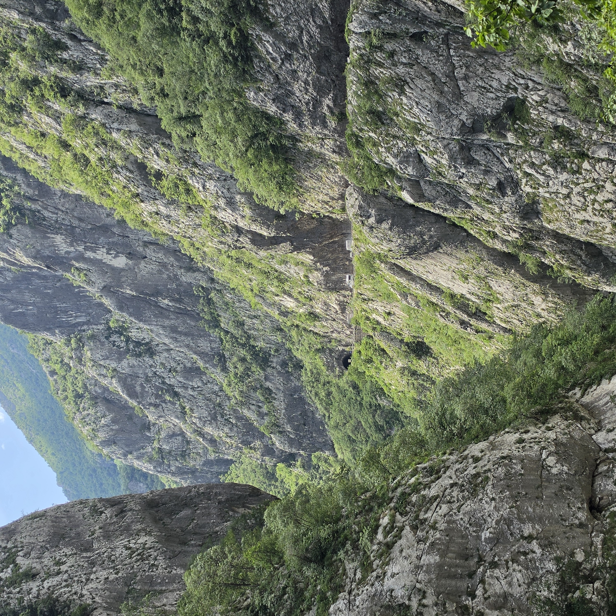 The image shows a view from below of a steep canyon or gorge with gray rock walls covered in patches of green vegetation. The image is taken in a mountainous area in the city of Vlahovići, Montenegro. A road with vehicles runs along one of the canyon walls, passing through a tunnel. The sky is visible in the upper left corner, and additional rocky terrain is visible in the foreground.