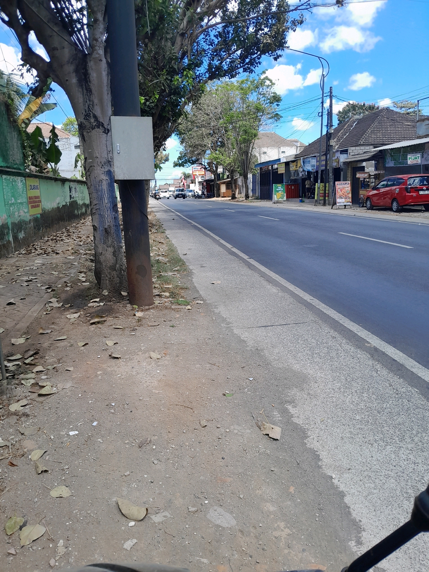 The image depicts a street scene in Talangagung, Indonesia. On the left side of the image is a sidewalk with a light green wall and some fallen leaves. A utility pole with a gray electrical box is situated between the sidewalk and the road. The road itself is paved and appears to be a two-lane street with traffic. On the right side of the road are several buildings, including shops and a red car parked in front of one of them. Signs for businesses like "ALTAMA" and "KABAMA" are visible. The sky is blue with some white clouds, suggesting it is daytime. A traffic light pole with wires is also visible in the distance. The overall impression is of a moderately busy street in a commercial area.