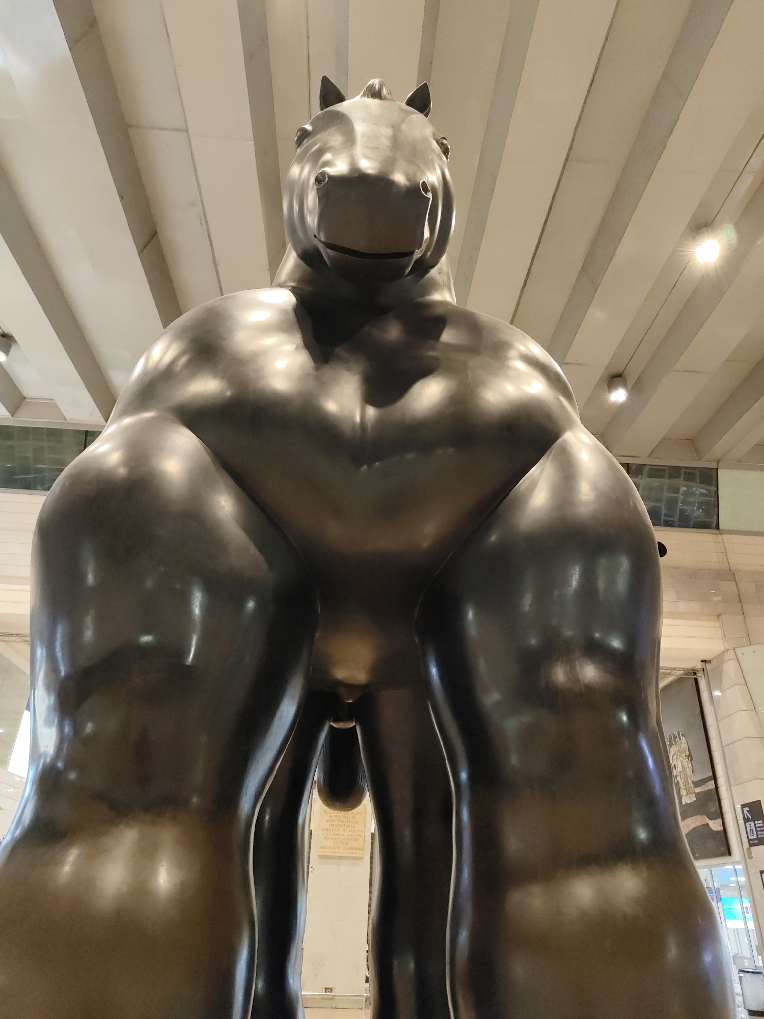 A low-angle, indoor shot showcases a bronze sculpture of a horse in El Prat de Llobregat, Spain. The statue's anatomy is exaggerated, emphasizing its muscular build. The horse is positioned centrally, viewed from a low angle that highlights its mass. The background features a high ceiling with parallel beams and artificial lighting. A partial view of a wall with signage and a mural is visible on the right side of the frame.
