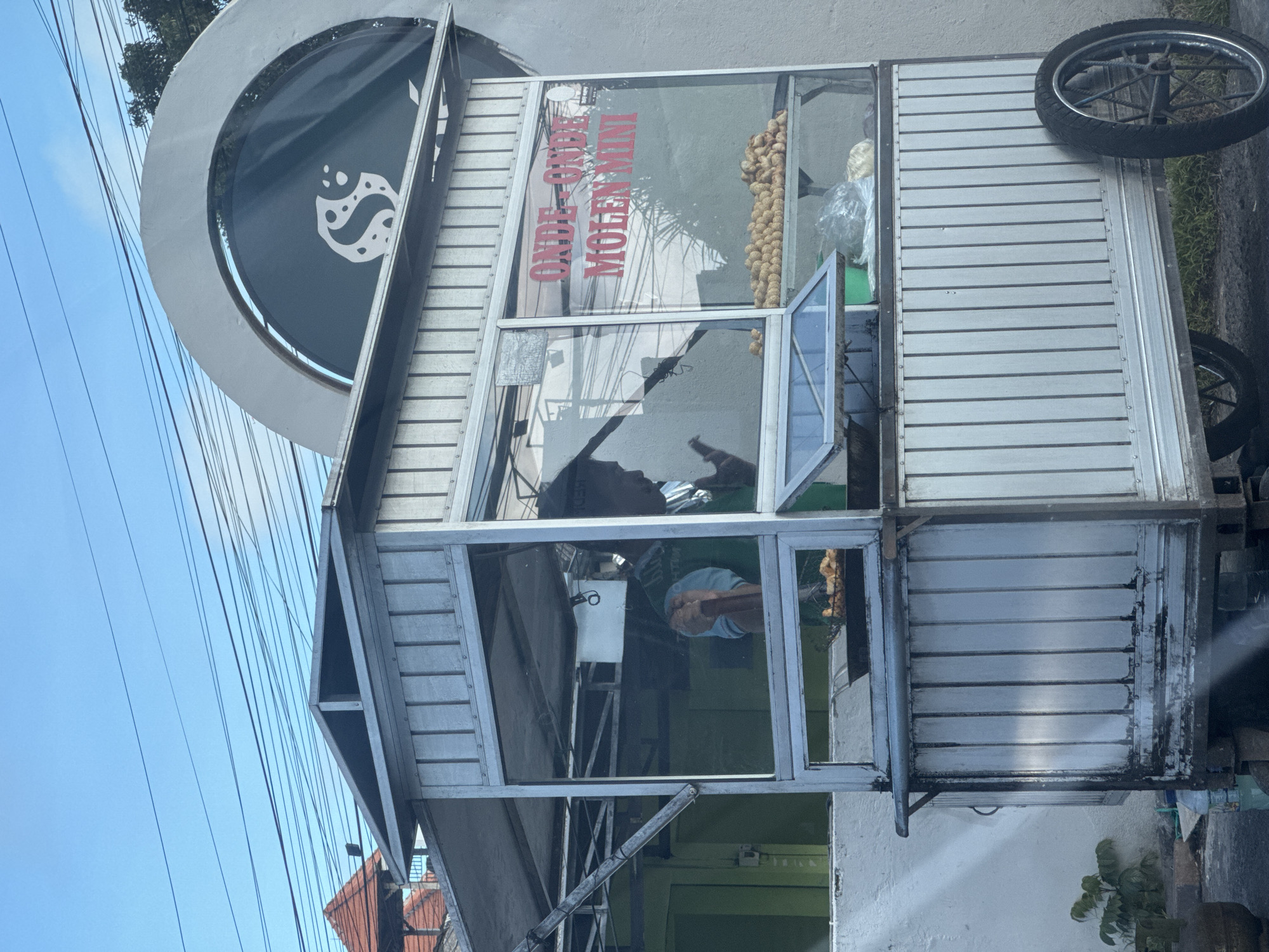 Here's a description of the image:
A mobile food stall in Kuta Selatan, Indonesia is visible. The stall is made of metal and glass, with "ONDE-ONDE MOLEN MINI" written on one of the glass panels. A sign with a circular logo is attached to the top of the stall. The stall is stocked with food. A person is visible inside the stall. The stall has wheels, indicating its mobility. Power lines are visible in the background. A building is visible in the background.