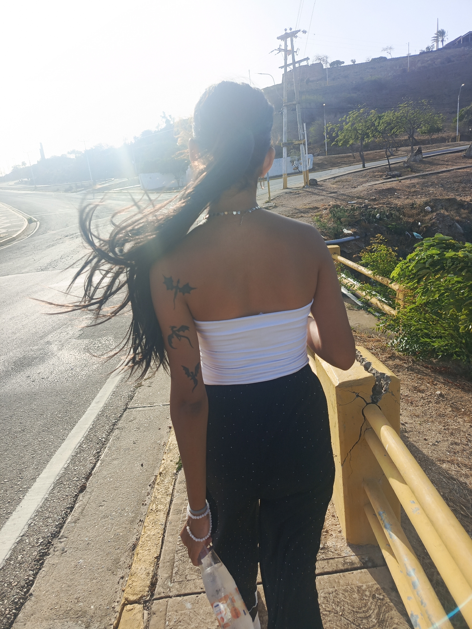The image shows a young woman from behind, walking outdoors on a paved surface, likely a bridge or pathway, with an asphalt road to her left. Her long, dark hair flows to her left, suggesting movement or a breeze. She is wearing a white strapless top and dark pants, possibly black with small white specks. Around her neck is a thin silver necklace with heart-shaped charms, and on her left wrist, she wears two light-colored beaded bracelets. Three dark tattoos, resembling dragons, are visible on her right upper arm and shoulder blade. She holds a plastic bottle in her right hand.
To the woman's right, a cracked and weathered yellow concrete barrier or railing runs alongside her path. Beyond this barrier, a dry, rocky hillside rises, featuring sparse green vegetation, a few trees, utility poles, and power lines. Another road or path can be seen winding up the hill. The scene is brightly illuminated by strong sunlight, causing a significant lens flare on the left side, indicating a clear, sunny day. The overall environment suggests an arid or semi-arid landscape, consistent with the location context of Juan Griego, Venezuela. No discernible text is visible on any objects.