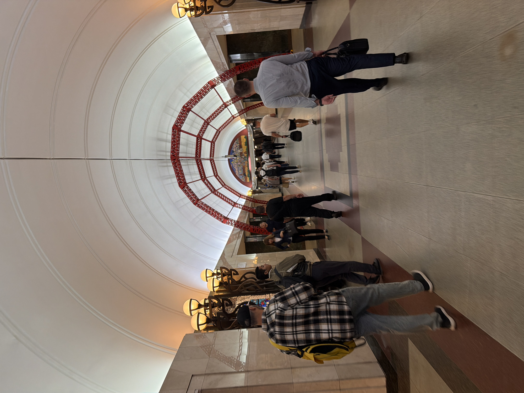 The image depicts a wide, ornate subway station tunnel with a high, arched ceiling. The ceiling is white with subtle curvature and decorative linear details. Red, ornate metalwork frames arch across the tunnel at intervals, supporting the ceiling structure and contributing to a grand aesthetic. Numerous people are walking in both directions along the polished floor, which features a patterned design of light and dark tiles with a distinct maroon border.
On the left side of the tunnel, decorative lamps with multiple spherical globes are mounted on intricately designed bases. The walls are clad in polished stone, with geometric patterns visible in some sections. Further down the tunnel, a large mural is visible on the far wall.
Several individuals are captured in motion. In the foreground, a person wearing a plaid shirt and jeans walks away from the camera, carrying a yellow and black backpack over their left shoulder. Another person in a light-colored shirt and dark pants walks towards the camera, carrying a black briefcase in their right hand. Other pedestrians, dressed in casual and business attire, are seen dispersed throughout the tunnel, some walking individually and others in small groups. The lighting in the station is warm and ambient, emanating from the decorative lamps and the tunnel ceiling.