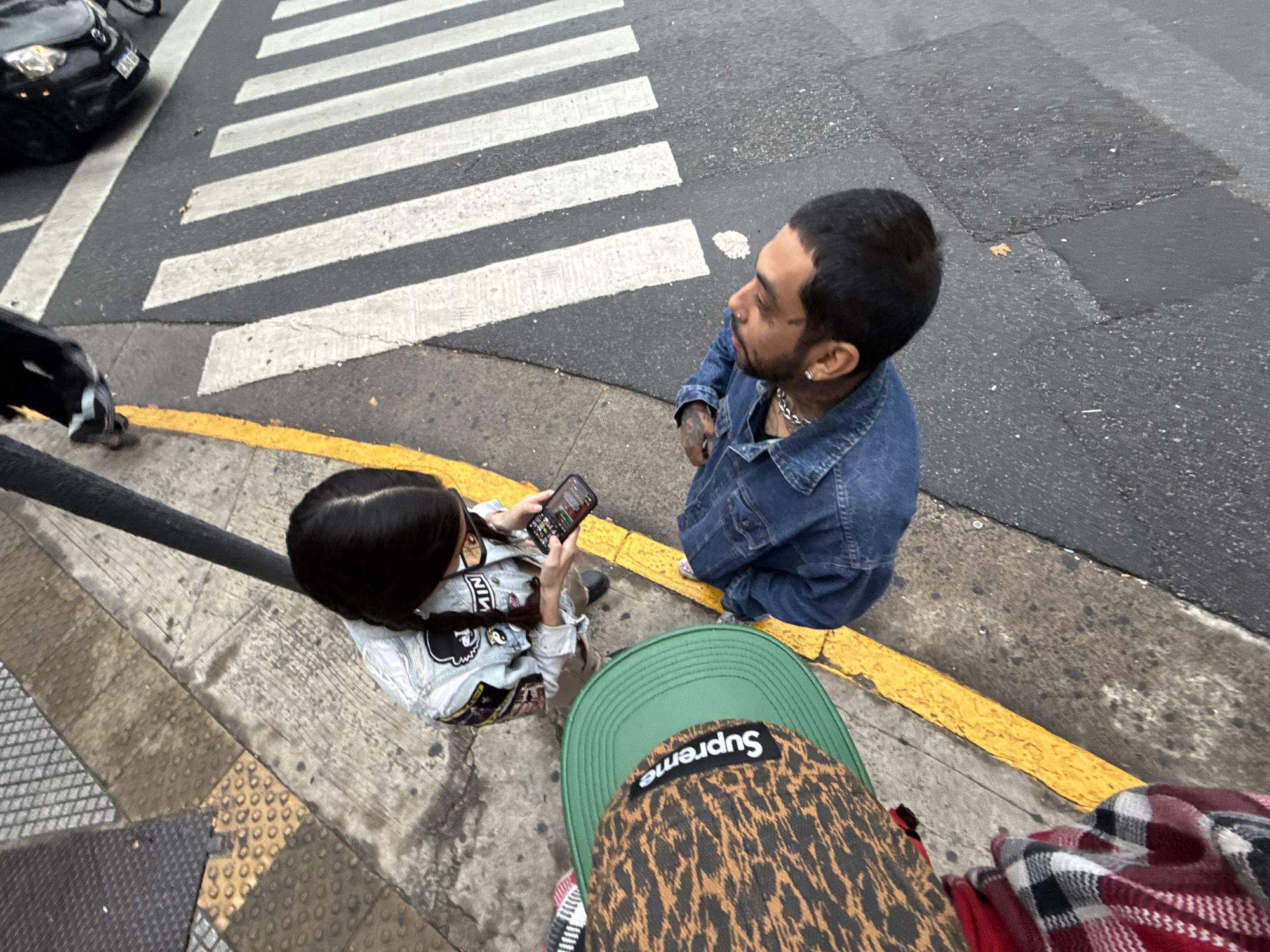 A high-angle urban street photograph from Buenos Aires, Argentina, captures two individuals standing on a sidewalk beside a crosswalk. In the mid-ground, a man with short dark hair, a beard, and facial tattoos (below his left eye) wears a denim jacket, a silver chain, and an earring. He looks upwards and to the left, observing something beyond the frame. Beside him, a woman with long dark braids holds and interacts with a smartphone, her gaze fixed on the screen. She wears a light-colored denim jacket featuring a Mickey Mouse silhouette patch and a "NIN" logo.
The foreground shows a green baseball cap with a leopard print brim, emblazoned with the inverted text "Supreme". Beside the cap, a red and white plaid fabric is partially visible. The setting includes a gray asphalt road with a prominent white zebra crossing. A black car is partially seen on the road in the upper left. A yellow curb line delineates the street from the sidewalk, which consists of varied concrete and tactile paving textures. The muted lighting suggests an overcast day or late afternoon, and the damp appearance of the street implies recent rain or cloudy weather. The scene conveys a casual, observational moment of city life.
