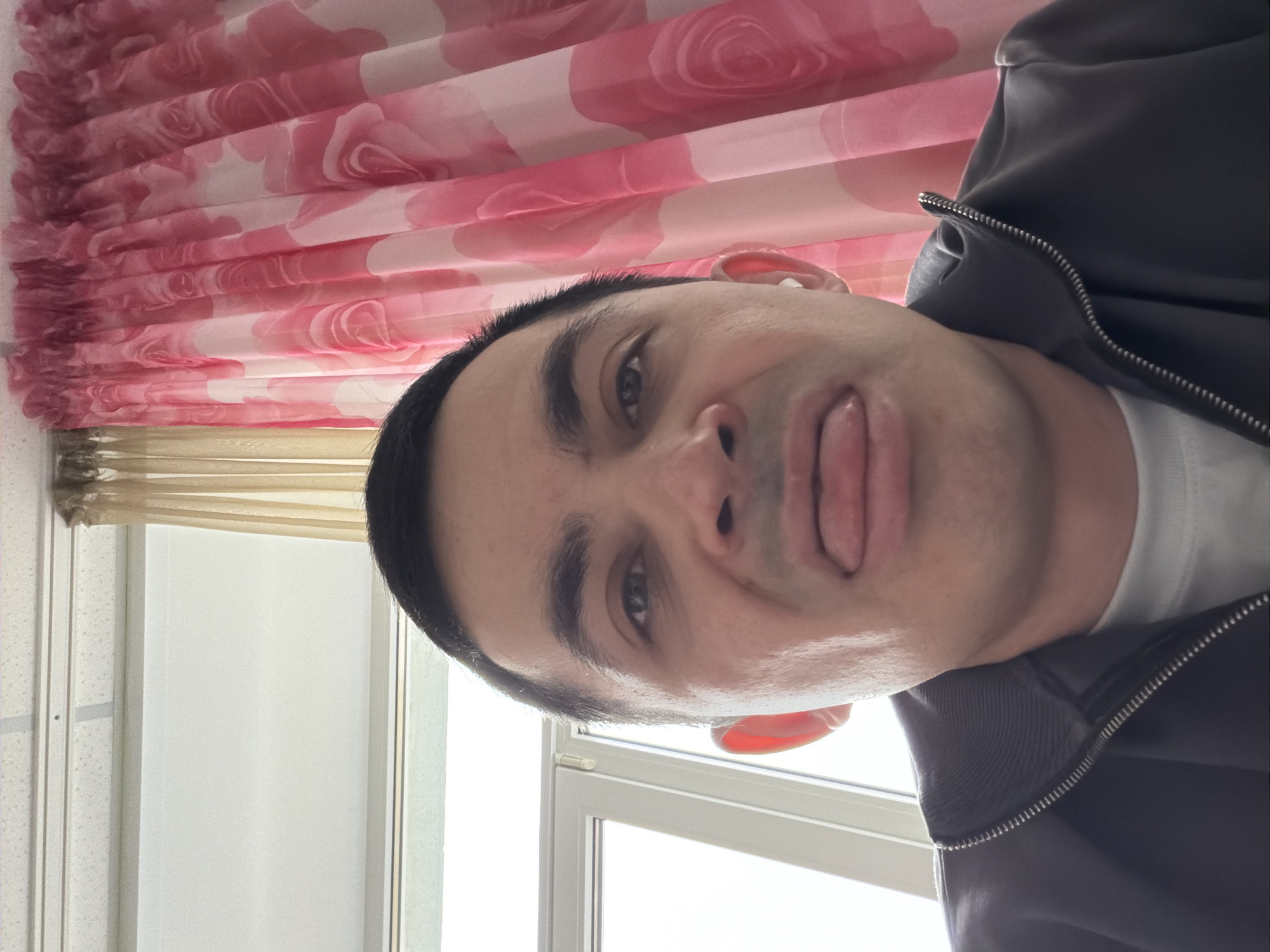 A man is depicted in a selfie, sticking his tongue out. He is wearing a white t-shirt and a dark gray jacket with a visible zipper. Behind him is a window, partially obscured by a beige blind and pink floral curtains. The location is not discernible from the image; therefore, it's impossible to confirm if it's in Izhevsk, Russia.