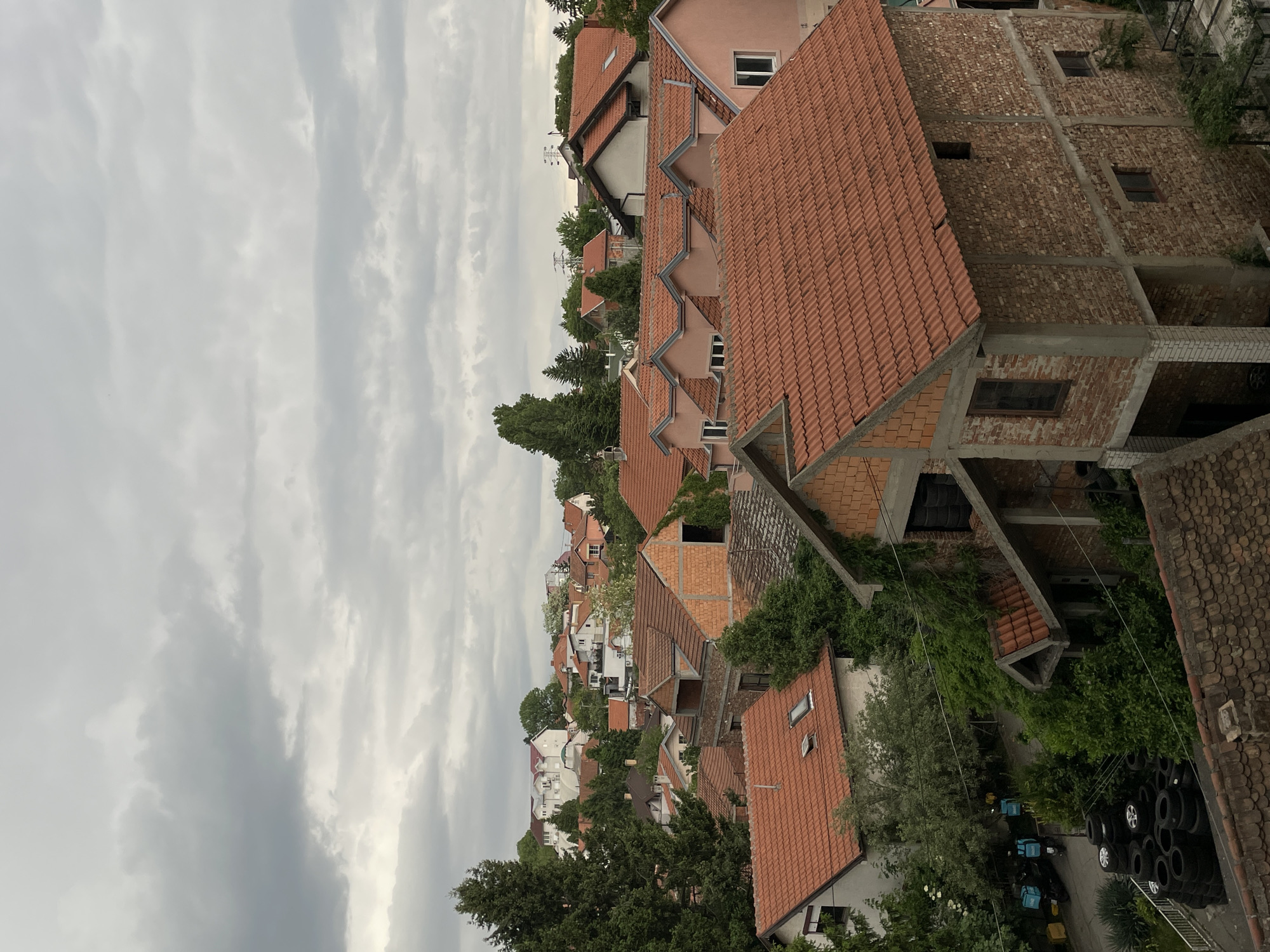 The image shows a residential area in Belgrade, Serbia, viewed from an elevated position. Numerous houses with red tile roofs are visible. Some buildings are under construction, with exposed brickwork and unfinished facades. Lush greenery is interspersed among the houses, including trees and climbing vines. In the foreground, a stack of tires is visible, along with delivery scooters and bicycles. The sky is overcast with cloudy.