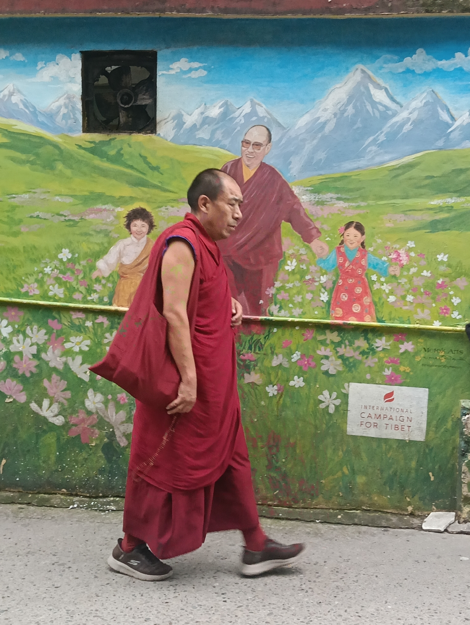 A man wearing a maroon robe walks past a mural in Dharamkot, India. The mural depicts a mountainous landscape with a blue sky and white clouds, a green field, and a variety of flowers. In the center of the mural, a figure resembling the Dalai Lama stands between two children, a boy and a girl. The man in the foreground is seen from the waist down, with his maroon robe trailing behind him. He is carrying a maroon bag over his shoulder and is wearing dark athletic shoes. A white sign with red lettering that reads "INTERNATIONAL CAMPAIGN FOR TIBET" is visible on the lower right side of the mural. The ground is a paved surface with visible pebbles.