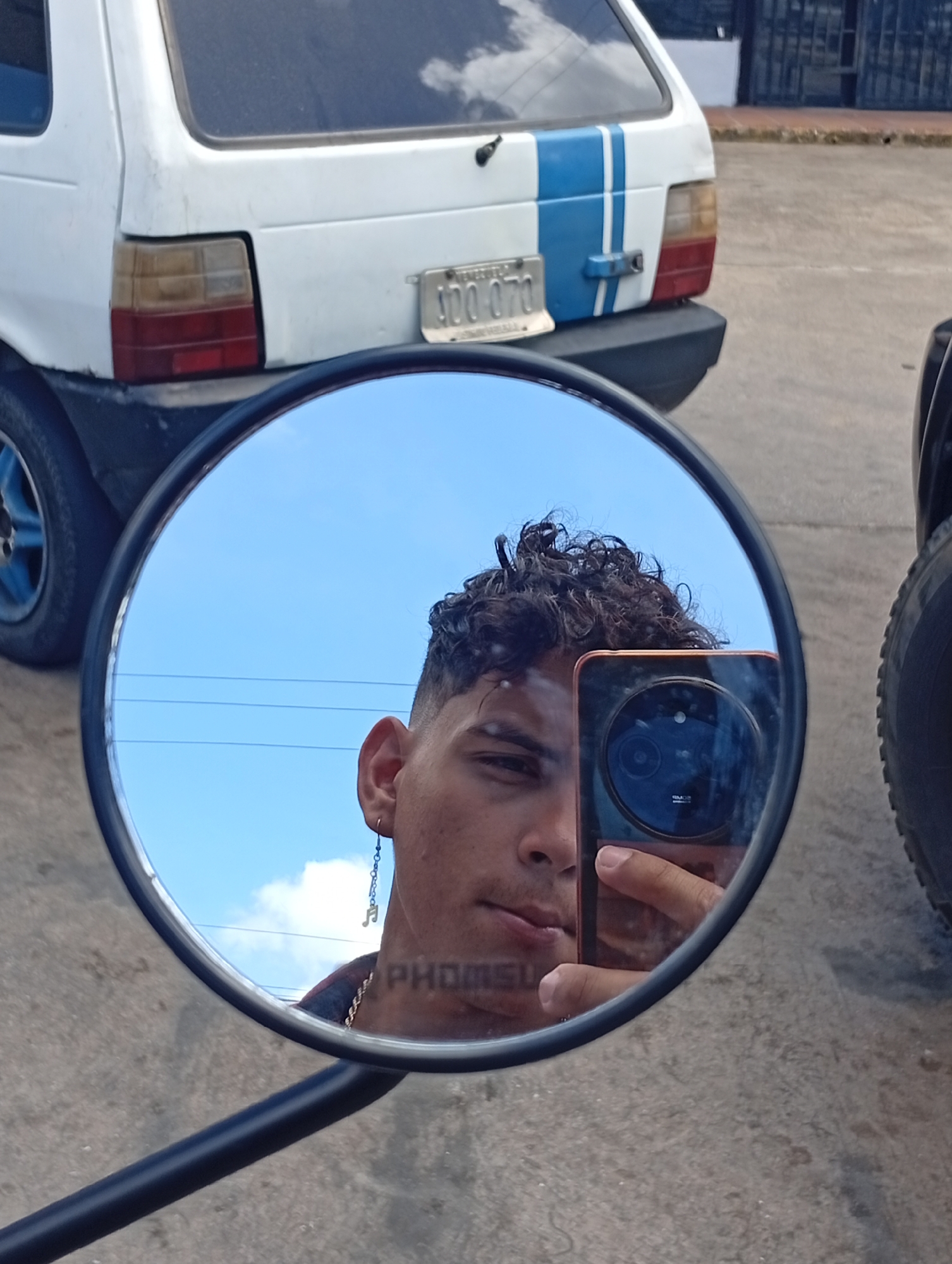 The image shows a person taking a selfie in a round motorcycle mirror. The reflection reveals the person's face, part of their phone, and the sky. In the background, outside of the mirror's reflection, there is a white car with a blue stripe and a license plate that appears to read "100 070". There is also a portion of a large black tire visible. A white wall or building can also be seen in the far background. Based on the presence of a car with a Venezuelan license plate and the description, the image was taken near Juan Griego, Venezuela. Further visual indicators for verification of this information would need to come from a larger area of the image.