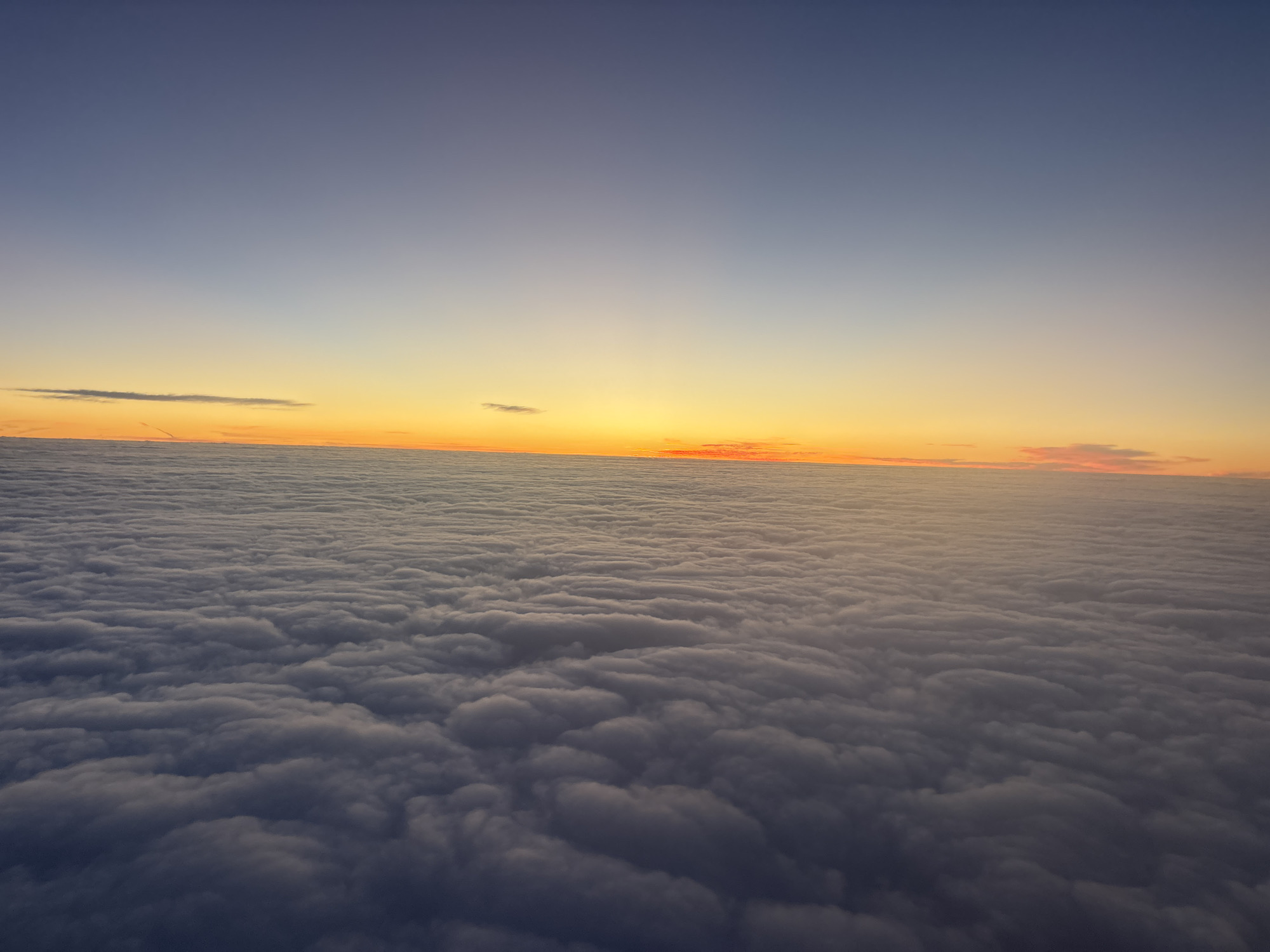 The image shows a landscape of clouds at sunset. The sky transitions from a deep blue at the top to a pale yellow-orange horizon. A dense blanket of clouds covers the lower portion of the image, appearing textured and somewhat turbulent. It is impossible to determine the location in Buenos Aires or any other city from this image. There are no people or other objects visible besides the sky and clouds.