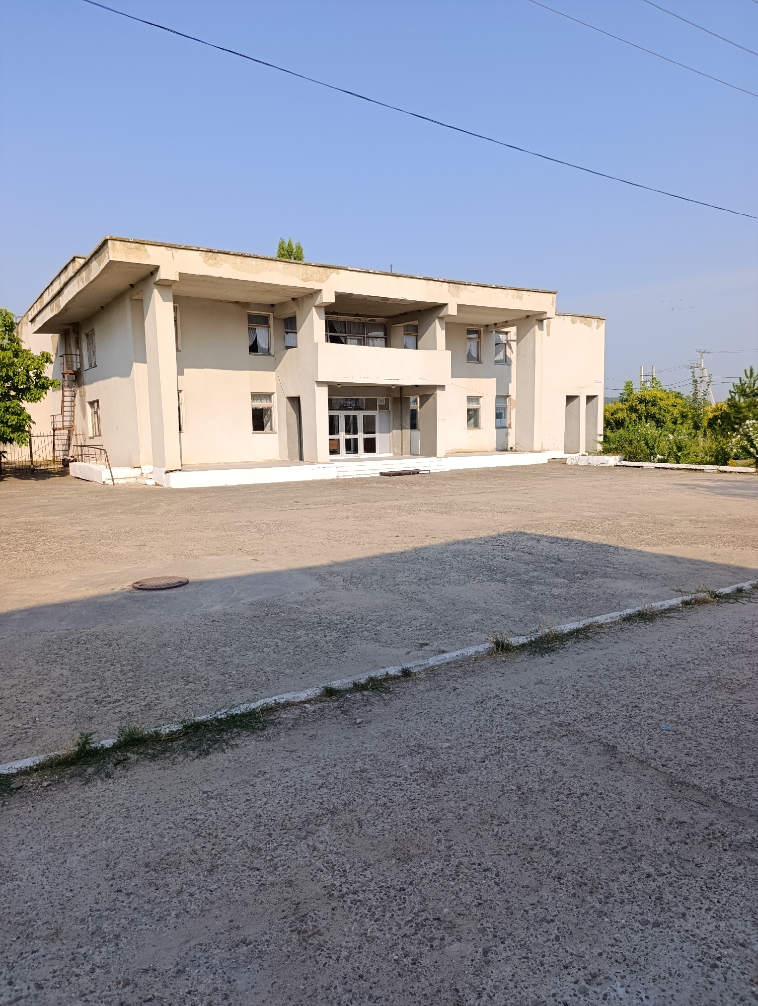 A wide shot captures a large, off-white, two-story building under a clear blue sky. The building has a flat roof and features multiple windows, some with curtains. A metal fire escape is visible on the left side. The ground in front of the building is a concrete-paved area, with a circular manhole cover in the foreground. The right side of the image shows lush green trees and vegetation. In the distance, power lines and poles are visible. The scene is static and appears to be an outdoor view of a building in Garmatskoye, Moldova.