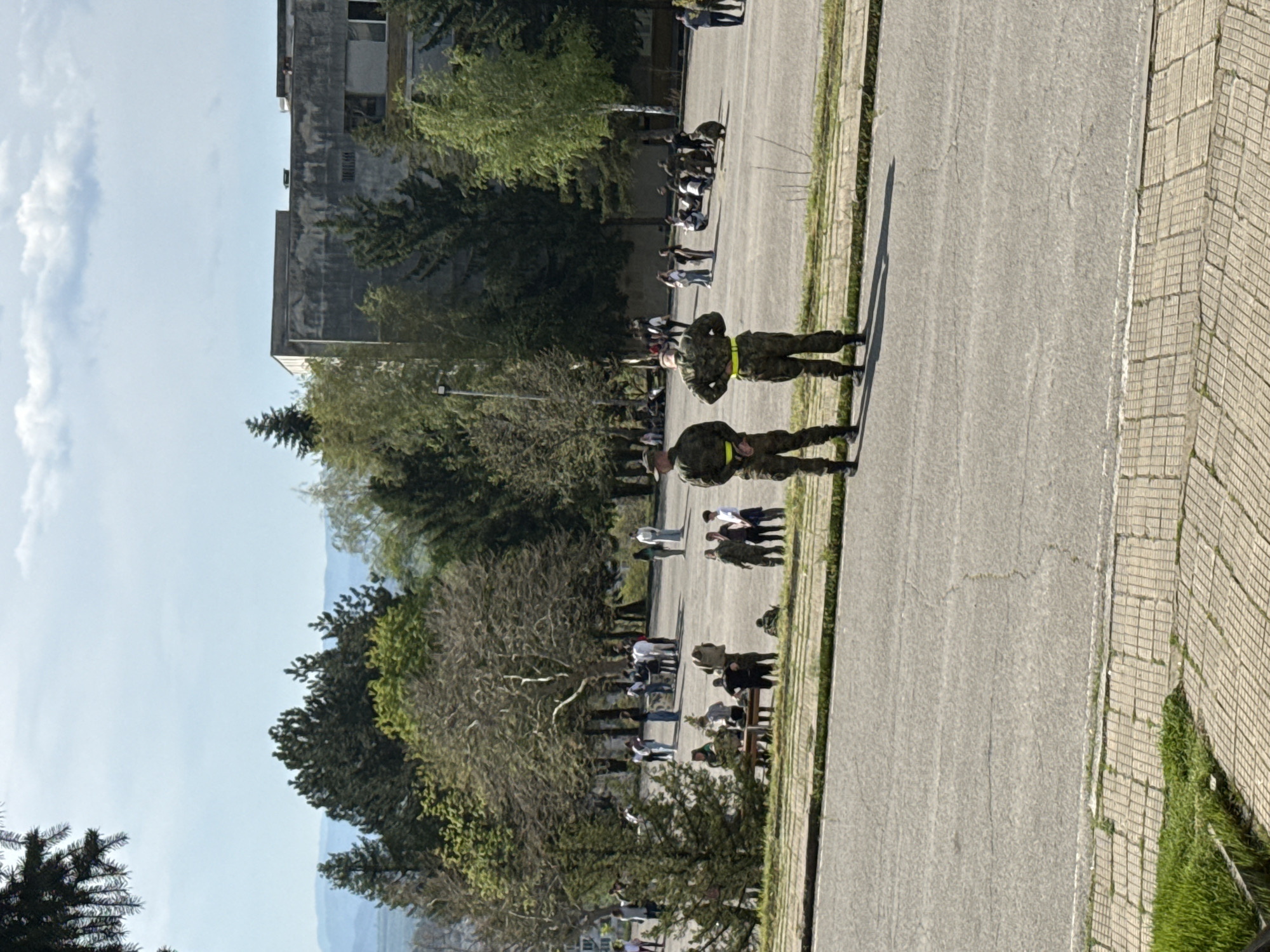 The image depicts an outdoor scene featuring people, a paved road, green spaces, and a building under a clear sky.
**Subjects:** Two individuals in military-style camouflage uniforms, augmented with yellow reflective belts, stand prominently on a paved road in the mid-ground. Their backs are mostly to the viewer, and they face towards the left side of the frame. Further back, positioned to the left of the road and partially obscured by trees, are several small groups of casually dressed people, some standing and others appearing to be seated. A large, multi-story, grey concrete building, exhibiting signs of age and weathering, dominates the background on the left. The area is rich with vegetation, including numerous trees with varying foliage density and green grassy patches along the edges of the paved surfaces. A wide paved road stretches horizontally across the frame, with a distinct patterned tiled sidewalk bordering its right side.
**Activity or event taking place:** The uniformed individuals appear to be standing guard or on patrol, maintaining a watchful stance. The other groups of people seem to be gathered casually, possibly observing their surroundings, socializing, or awaiting an unidentifiable event. The overall atmosphere suggests a routine outdoor assembly or presence.
**Notable details:** The time of day is likely late morning or early afternoon, indicated by the bright sunlight and relatively short shadows cast by the uniformed figures. The weather is fair, with a clear blue sky dotted by a few scattered white clouds. Location cues, such as the military attire and the utilitarian architecture of the background building, suggest a setting that could be a military base, a public campus, or a similar institutional environment. Distant hills or mountains are faintly visible on the horizon in the far background. No visible text is present in the image.