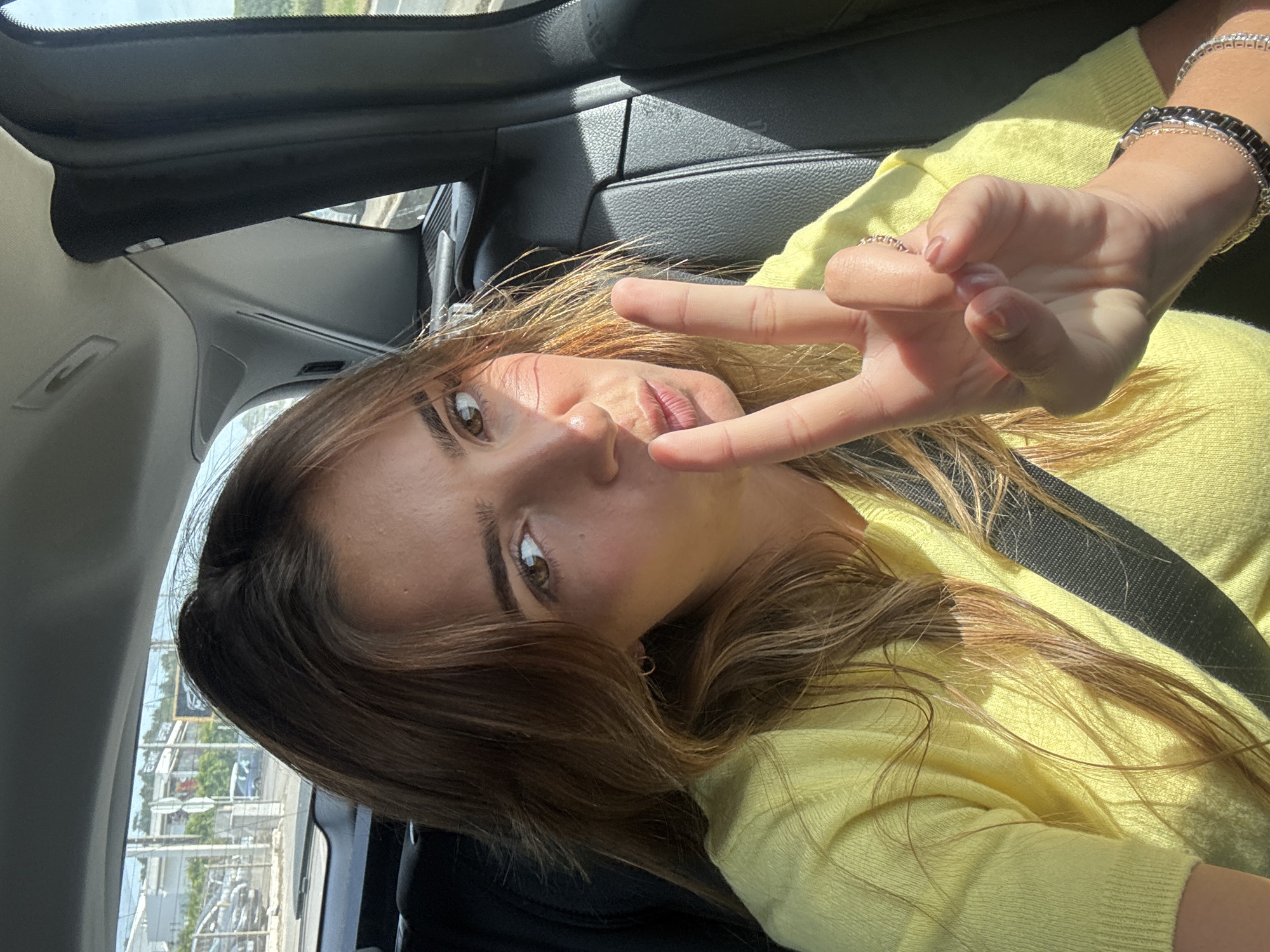 The image captures a young woman inside a car, likely in Torres Vedras, Portugal, taking a selfie or being photographed from an elevated angle. She lies back with her head tilted, looking directly at the camera with a relaxed expression and slightly pursed lips. Her long, brown hair, with visible lighter streaks, cascades around her face and over her bright yellow long-sleeved top or sweater.
Her right hand is raised, forming a "peace sign" or "victory sign" with her index and middle fingers extended. On her right wrist, she wears multiple bracelets, including a silver-toned chain and a black-and-silver link design. A ring is visible on her left ring finger. A dark seatbelt diagonally crosses her chest.
The car's interior features dark upholstery and trim. Through the windows, a bright, blurry outdoor scene is visible, suggesting an urban or semi-urban landscape with buildings and utility poles, illuminated by strong sunlight. The bright lighting indicates it is daytime and sunny. No visible text appears in the image.