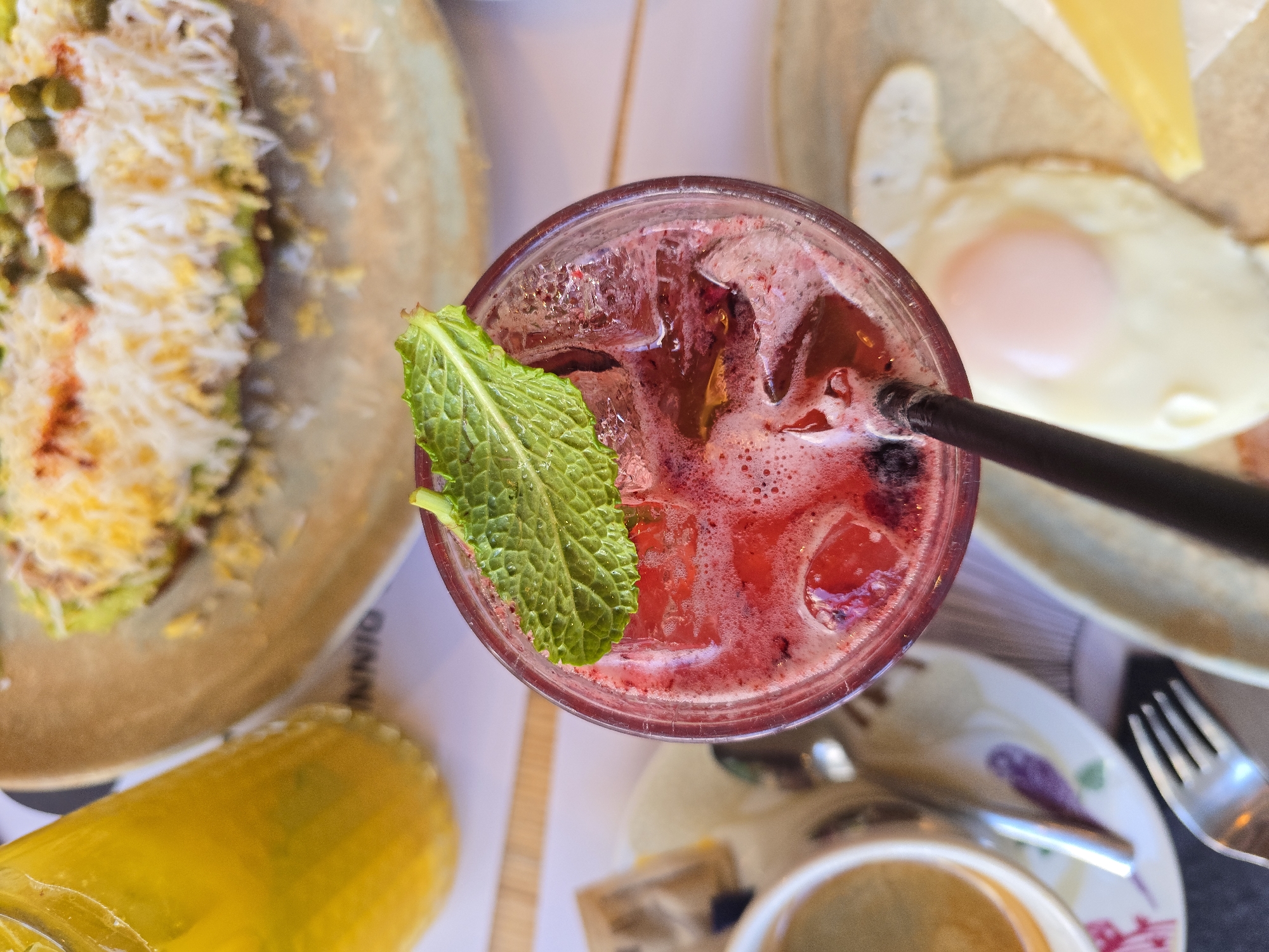 This overhead shot presents a diverse spread of food and beverages laid out on a table at a cafe or restaurant in Varna, Bulgaria. The central item is a glass of vibrant red or purple liquid, likely a berry drink or cocktail, filled with ice cubes and garnished with a fresh green mint leaf. A black straw protrudes from the drink.
To the upper left, a light brown plate holds a dish adorned with shredded white ingredients, possibly cheese or a similar topping, interspersed with green elements like capers and traces of a yellow or orange sauce. Part of the word "INNIO" is visible on a placemat or plate design beneath this dish.
In the upper right, another dish features a neatly cooked fried egg with a pale, soft-looking yolk, presented alongside a solid slice of yellow cheese. To the bottom left, a tall, yellow-orange bottle or glass, possibly containing juice, is partially visible. The lower right area reveals a white coffee cup on a saucer with a distinct floral pattern, accompanied by a spoon and a metal fork, suggesting a complete place setting. The scene captures a daytime dining experience.