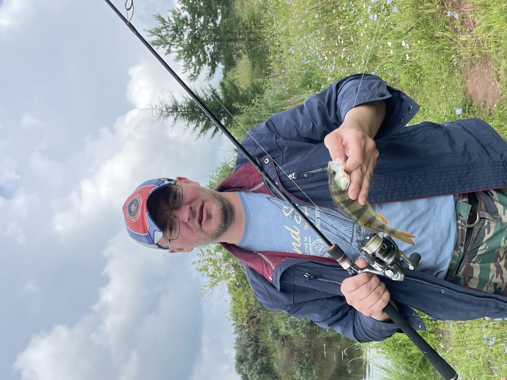 A medium shot captures a man holding a small fish and a fishing rod on a cloudy day. The man, wearing glasses and a baseball cap, faces the camera with an open mouth, suggesting he is speaking. He is dressed in a blue jacket over a t-shirt and camouflage pants. The fishing rod, with a reel, extends from the right side of the frame. The background features a body of water on the left and a field of wildflowers and trees on the right, indicating a rural or natural setting. The specific location, the city of Кумёны, Russia, places this scene within a region known for its natural landscapes and fishing opportunities.