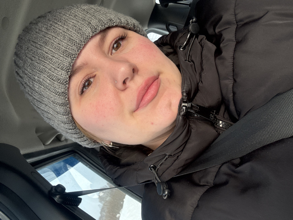 The scene depicts a female individual inside a vehicle. As per the query, the location is the city of Laiga, Russia, though no specific geographical identifiers are visible in the image itself. The individual, appearing to be an adult, is oriented towards the camera, displaying a neutral expression. She wears a grey knitted beanie and a black puffer jacket with a visible front zipper and drawstring collar details. A black vehicle seatbelt is positioned diagonally across her chest and left shoulder. Her visible facial features include light skin with areas of redness on the cheeks, brown eyes with distinct eyelashes, and pink-toned lips. A small, light-colored stud piercing is present on her left nostril, and a silver-colored hoop earring is visible on her left ear. Through the vehicle's left-side window, a blurred outdoor environment is visible, characterized by snow-covered ground and what appears to be bare vegetation, suggesting a winter setting.