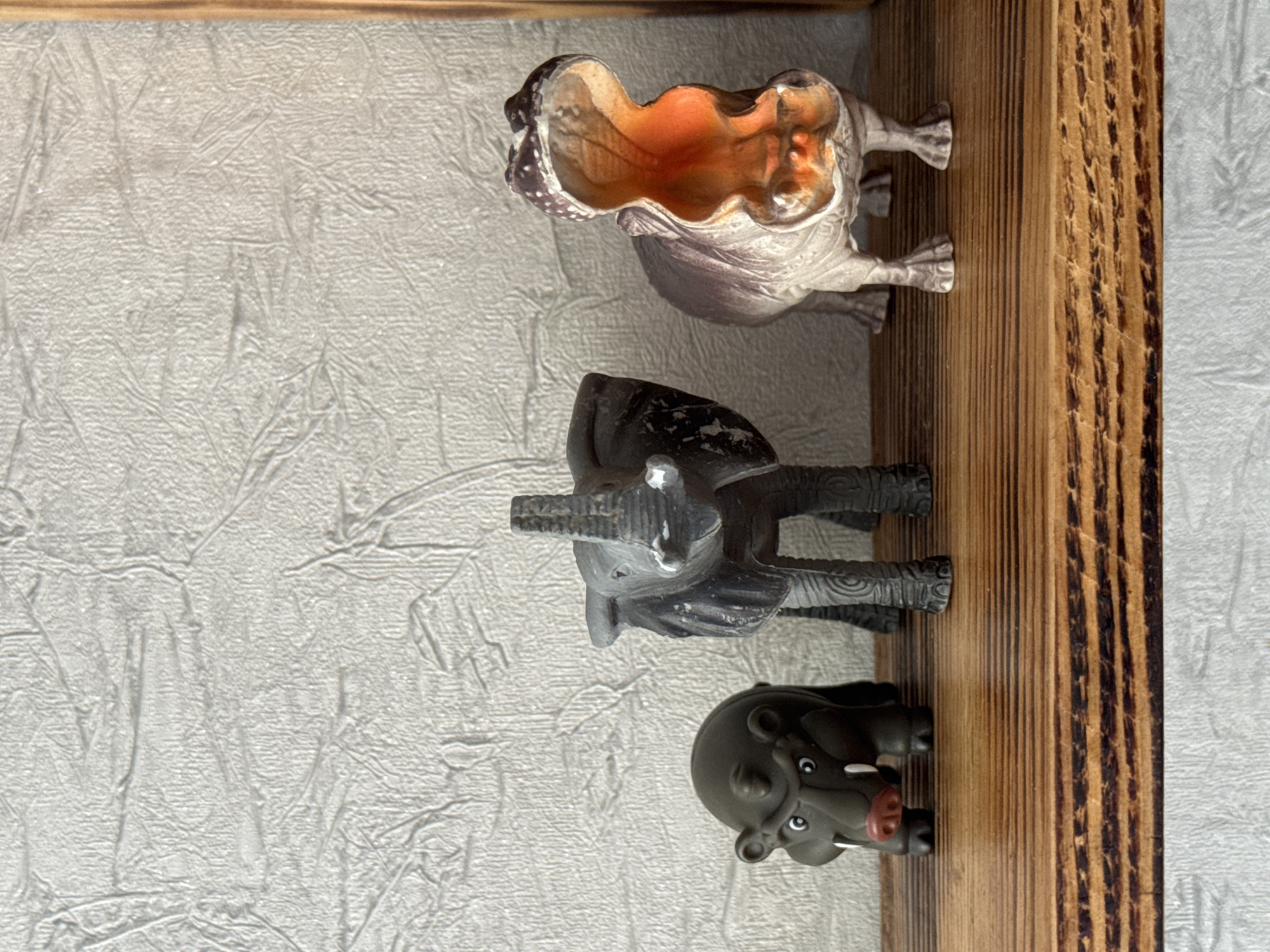 A close-up, eye-level shot shows three toy animals lined up on a wooden shelf against a textured, light-colored wall. The animals, from left to right, are a small, dark gray hippo with a red nose, an elephant with its trunk raised, and a large, light brown and purple hippopotamus with its mouth wide open revealing an orange interior. The shelf has a visible wood grain pattern. The background wall has a rough, stucco-like texture. This scene is located in the Яромінскі сельскі Савет, Belarus.