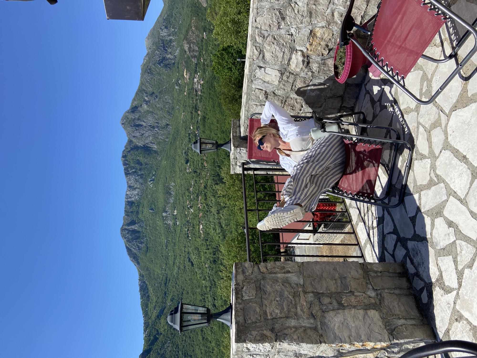 The image depicts a woman relaxing in a red lounge chair on a stone patio, likely in Sotonići, Montenegro, given the mountainous landscape. She is wearing a white jacket, light-colored top, vertically striped pants, and white sneakers. Sunglasses rest on her face, and her hands are behind her head. A stone wall surrounds the patio, with a metal railing in front of the woman. Beyond the railing, a steep, green mountain dominates the background, set against a clear, blue sky. A decorative lamppost stands near the wall, adding to the ambiance. Another red lounge chair is partially visible in the upper right corner of the image. The ground of the patio appears to be paved with uneven stone tiles.