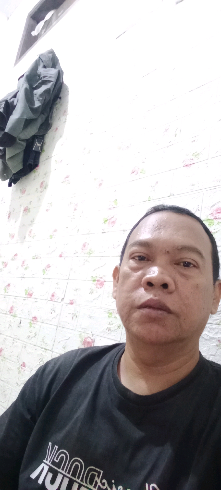 The image is a selfie taken by a middle-aged man with brown skin and short, dark hair. He is wearing a dark t-shirt with white text that is partially obscured. He is looking directly at the camera with a neutral expression. The background consists of a wall with a floral pattern and a gray jacket hanging on the left side of the frame. The lighting is bright, suggesting an indoor setting with artificial light. The scene appears to be a bedroom or a casual living space. The quality of the image is moderate, with some graininess and slight blur. There is no discernible information in the image that would indicate a specific location within Tasikmalaya, Indonesia.