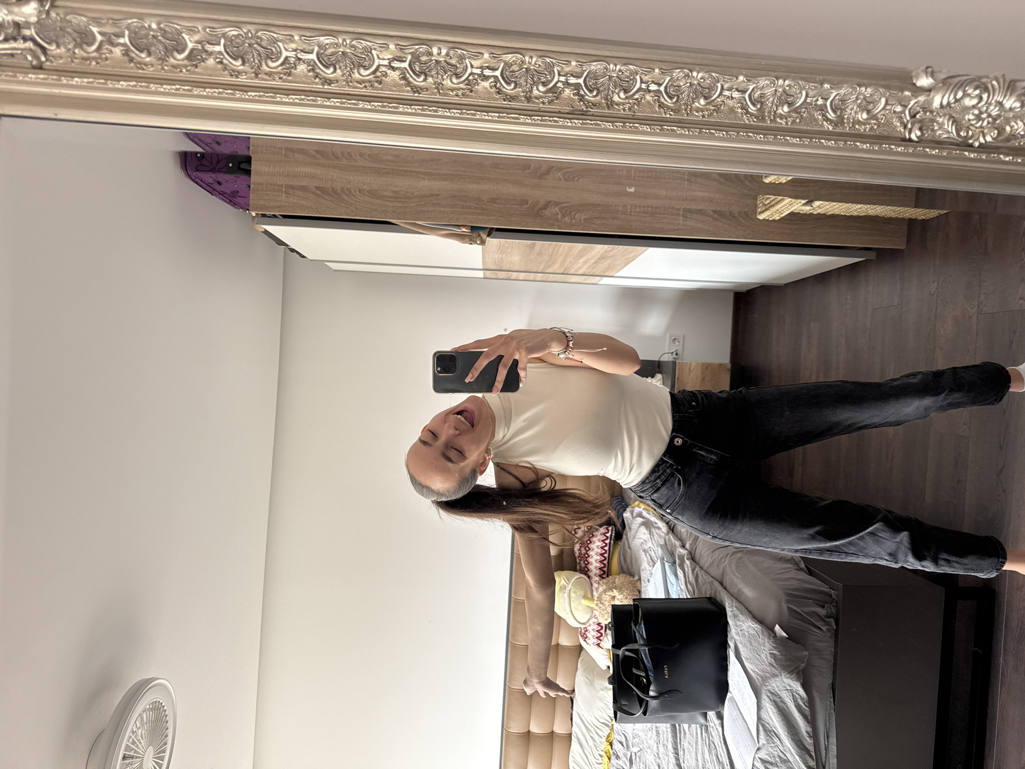 A woman is taking a selfie in a mirror. The mirror has an ornate silver frame. The woman is wearing a white sleeveless top and black jeans. She is holding a smartphone in her right hand. She has long brown hair pulled back in a ponytail. She is smiling with her mouth open. The room appears to be a bedroom with a bed, a headboard, and a nightstand with a black purse on it. There is also a white wall with a circular fan mounted on it, and a wardrobe. The location is likely a private residence in Sofia, Bulgaria.