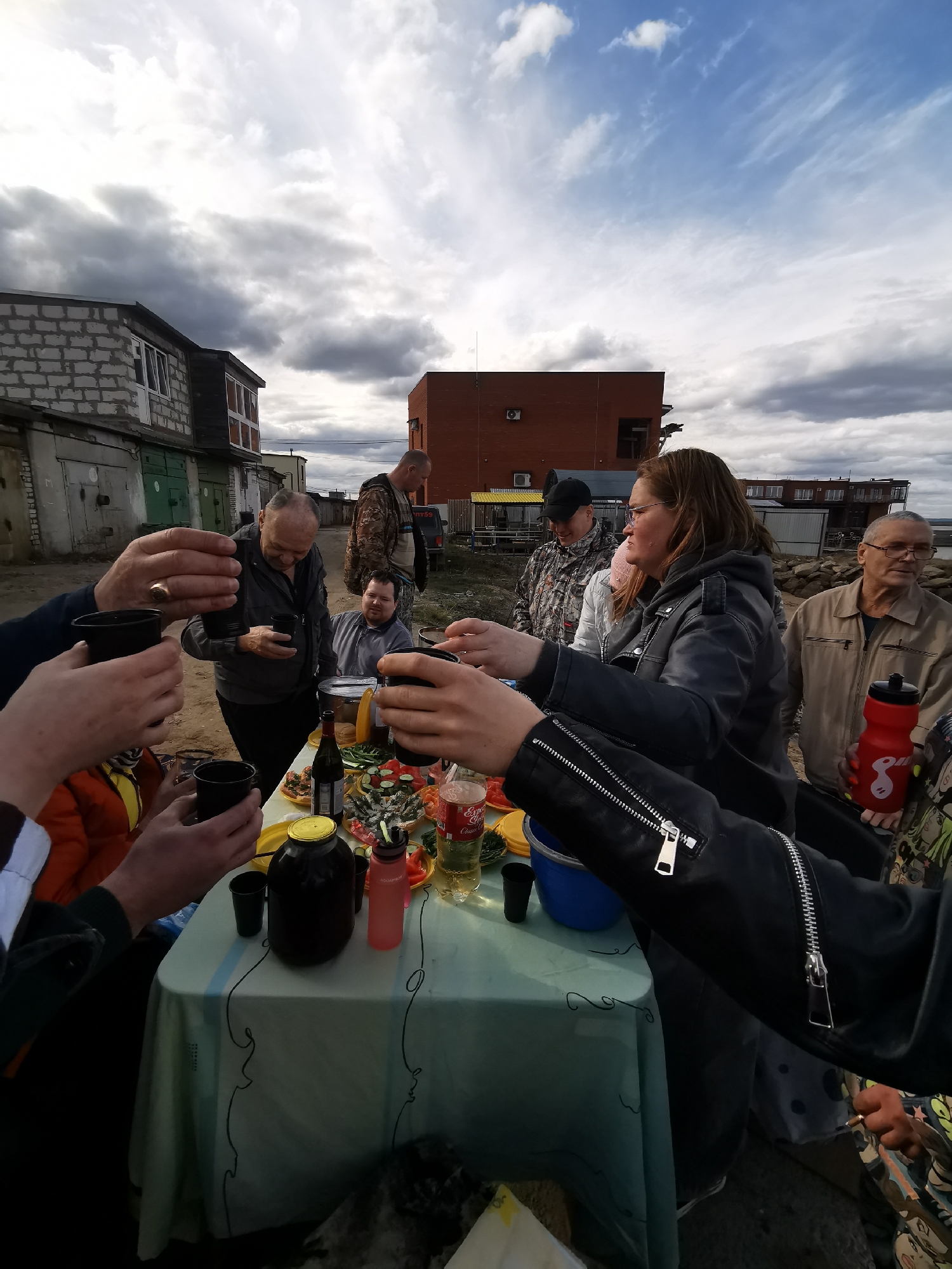 In the city of Unknown, Unknown, a group of individuals is gathered around a table laden with food and drinks. The individuals are holding black cups, seemingly engaged in a toast. The table is covered with a light-colored tablecloth adorned with dark, swirling designs. On the table, there are various items, including a bottle of wine, a large jar with a gold lid, and a red water bottle. Several people are standing around the table, some holding their cups aloft in a toasting gesture. A building constructed from cinder blocks and another building made of red bricks can be seen in the background. The sky overhead is filled with dark, ominous clouds.
