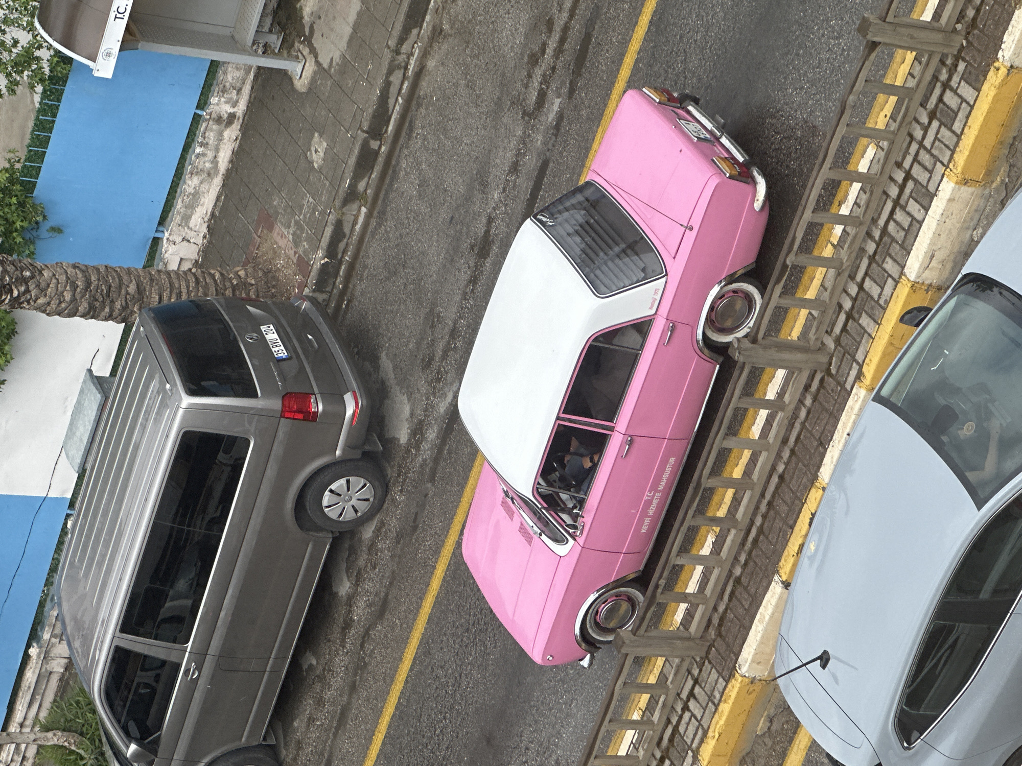 The image presents an aerial view in Fethiye, Turkey. A gray multi-purpose vehicle (MPV) with a license plate "35 BTU 204" is parked alongside a road. Adjacent to it, a vintage pink and white car, possibly a classic model, is parked alongside a curb. This car features a license plate and lettering reading "TC, KEYFI HIZMETE MAHSUSTUR." To the right of the pink car, a light gray car is partially visible. The environment shows a mix of asphalt road, painted curbs, and hints of nearby buildings. A palm tree is visible near the MPV.