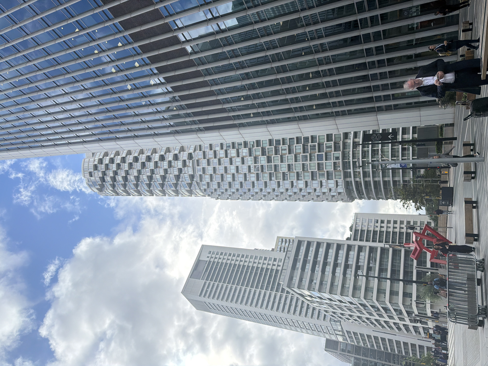 This image captures a vibrant, modern urban landscape in London, United Kingdom, characterized by striking contemporary architecture under a bright, cloudy sky.
The scene is dominated by several high-rise buildings. On the left, a tall, glass-fronted skyscraper reflects the blue sky and white clouds, featuring a facade composed of horizontal rows of reflective windows and dark grey panels. Adjacent to it, a distinctive, cylindrical building stands with a textured facade made up of numerous small, square-like windows arranged in an intricate pattern. Further back and to the right, several other modern, light-colored buildings with multiple windows and balconies contribute to the dense urban environment.
At ground level, several people are visible. One man, prominently featured in the middle-right foreground, is dressed in a dark suit and white shirt, holding an object, possibly a phone. Other individuals are scattered across the scene, some walking, some standing, suggesting routine daily activity. Metal crowd barriers are present on the ground, along with what appear to be street furniture like benches. A notable red, abstract sculpture, formed from interlocking geometric shapes, adds an artistic element to the urban space on the right side.
The time of day appears to be daytime, with clear visibility and ample natural light illuminating the buildings and surroundings. The weather is partly cloudy, with a mix of blue sky and scattered white cumulus clouds. A street sign with a left-pointing arrow and a pedestrian/trolley symbol is visible, indicating direction. The overall impression is one of a bustling, architecturally rich city environment.