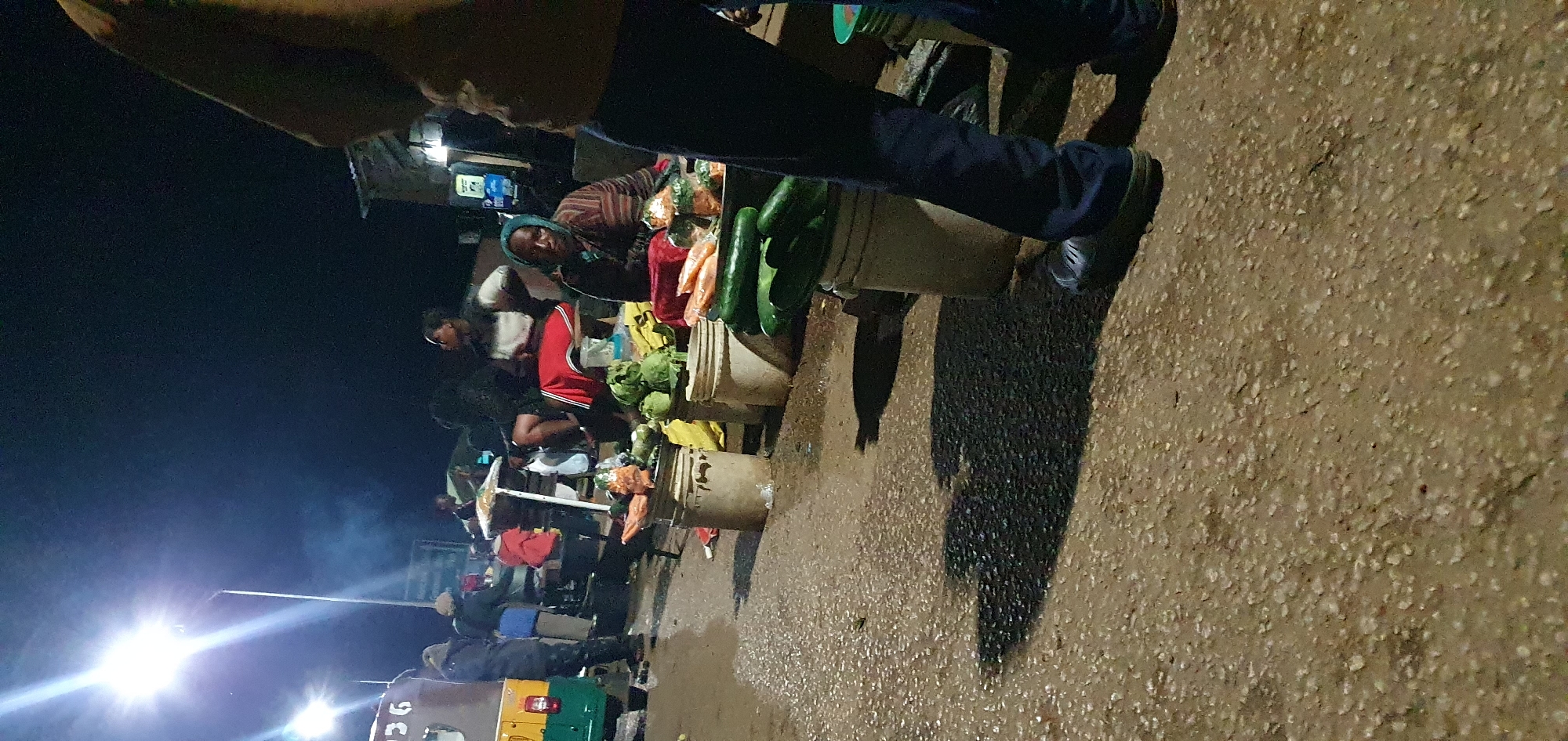 This is a nighttime street market scene in Bukuru, Nigeria. A brightly lit street lamp illuminates the area, casting long shadows. A vendor, wearing a headscarf and a striped patterned top, sits behind a display of produce, which includes large green vegetables that appear to be cucumbers and other leafy greens, as well as some orange items that might be carrots or sweet potatoes. Several large plastic buckets are used to hold the vegetables. In the background, other market-goers and vendors are visible, with a three-wheeled vehicle parked to the left. The ground is paved and appears to be wet, reflecting the light. The overall atmosphere suggests a busy, ongoing market transaction despite the late hour.