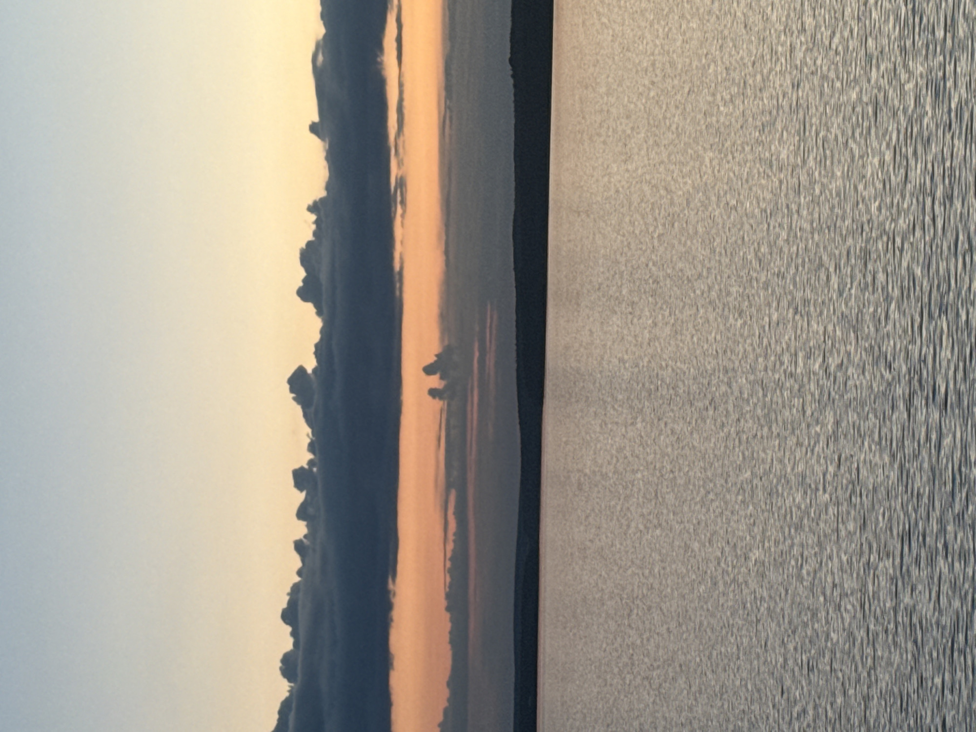 The image presents a serene and moody landscape, likely captured during either sunrise or sunset, over a large body of water in Votkinsk, Russia. The scene is divided horizontally into three main sections: the sky, a distant landmass, and the water.
The upper portion of the frame is dominated by the sky, which exhibits a soft gradient from a muted, light blue-gray at the top to warm, gentle orange and yellow tones closer to the horizon. Along the horizon, a distinct and somewhat dramatic band of dark, heavy clouds stretches across the scene, forming an uneven, silhouetted edge against the brighter sky. Below these clouds, a thin, dark line signifies a distant shoreline or landmass, likely covered with trees, appearing as a silhouette.
The lower half of the image is filled by the textured surface of a body of water, possibly a lake or reservoir. The water's surface is covered in small, shimmering ripples, reflecting the ambient light and creating a detailed, almost abstract pattern. The ripples appear more pronounced and linear towards the bottom right of the frame. The overall light suggests a calm, early morning or late evening atmosphere. No people or visible text are present in the image.