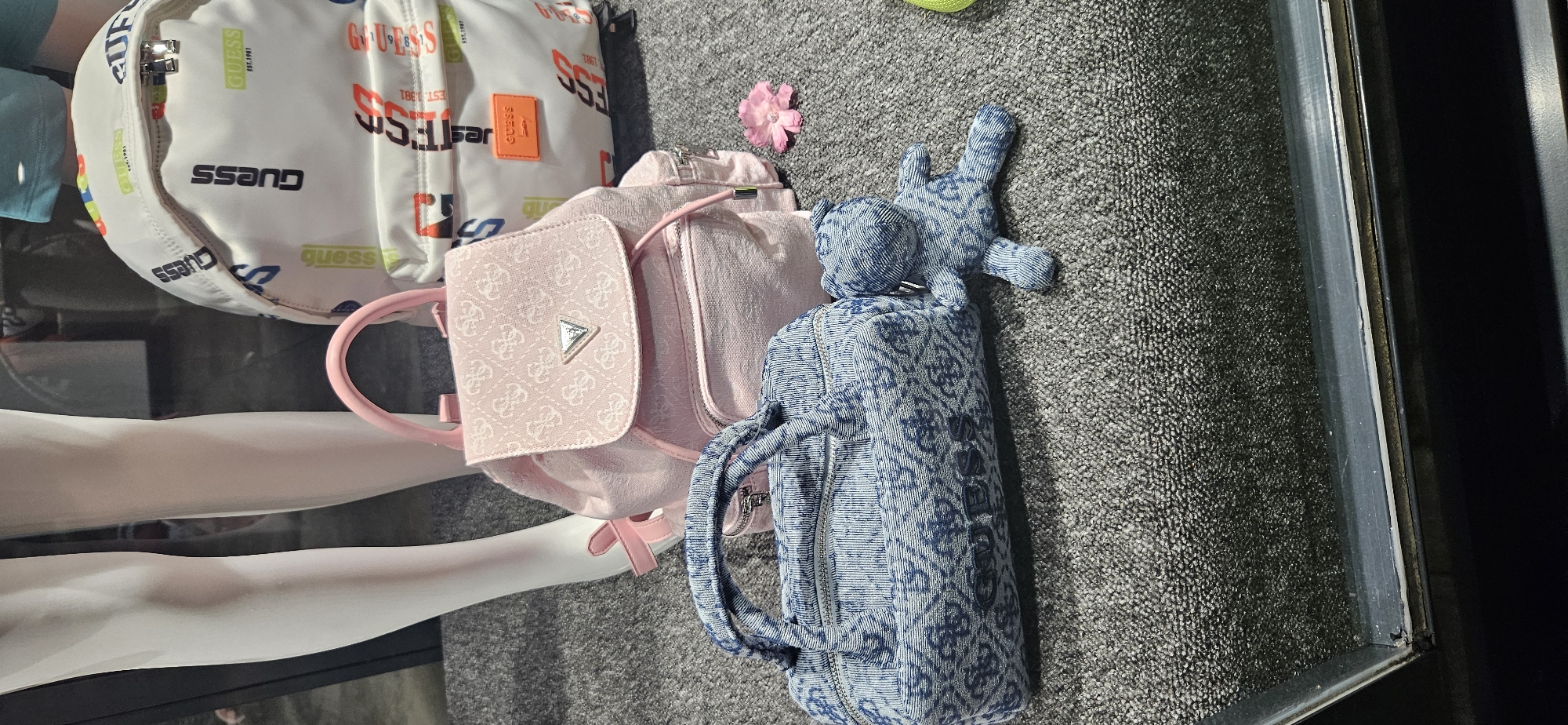 The image depicts a retail display of Guess brand accessories. In the foreground, a denim-patterned duffel bag with the "GUESS" logo is placed on a grey carpeted surface. To the right of the duffel bag is a small, denim-patterned teddy bear. Behind the duffel bag, a pink, patterned backpack with a triangular Guess logo is positioned. To the right of the pink backpack is a larger white backpack with a repeating Guess logo pattern in various colors. A single pink flower rests on the carpet between the pink backpack and the duffel bag. In the background, a mannequin's legs, clad in white stockings, are visible. The display appears to be in a shop window, with a dark frame at the bottom of the image suggesting a glass barrier. The overall setting is indoors, likely a clothing or accessory store. The city of Hévíz, Hungary is not visible or suggested by any elements within the image.