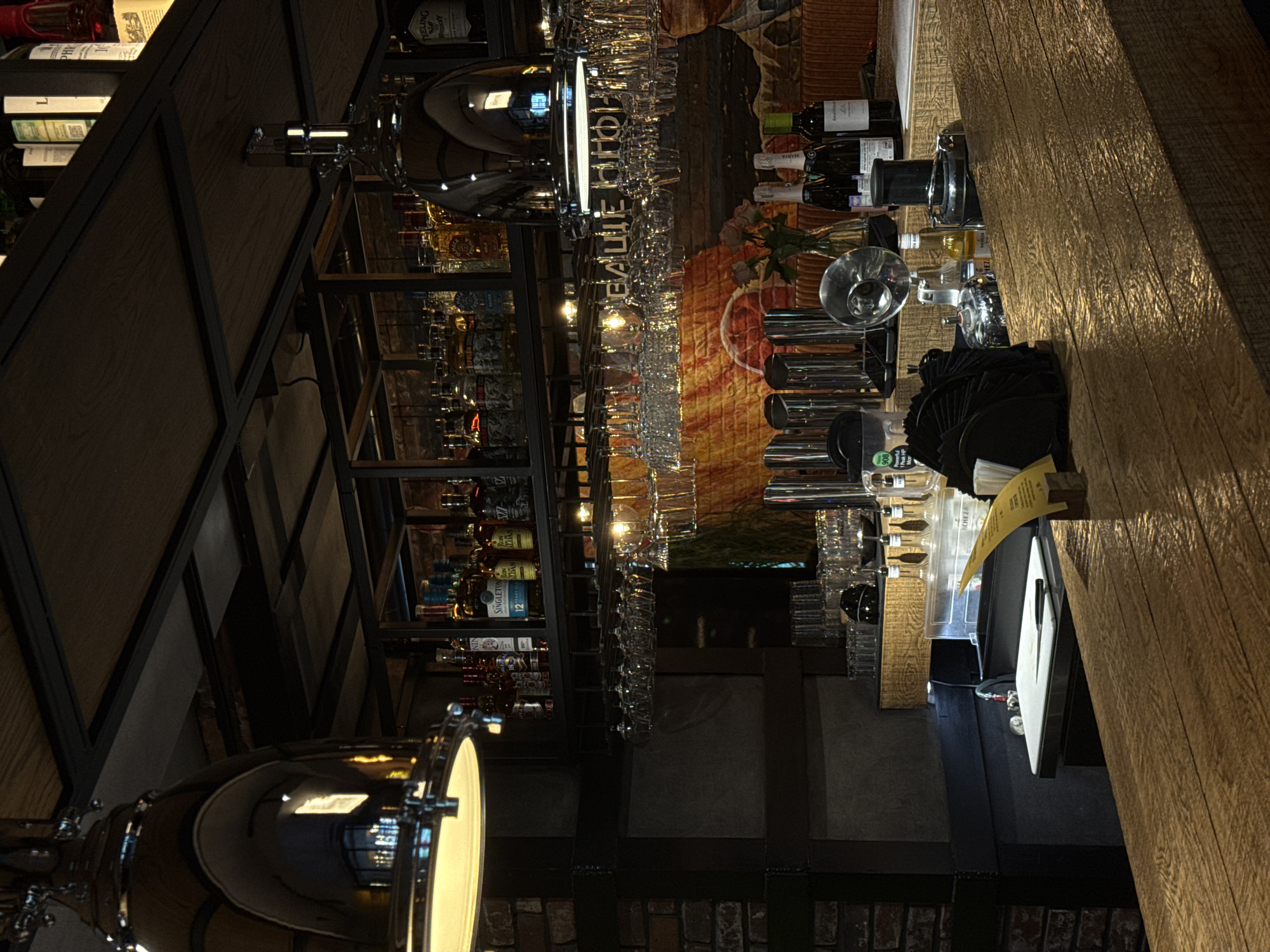 The image depicts an indoor bar area in Izhevsk, Russia, identified by the visible Russian word "БАШНЯ" (Bashnya) on a wall mural, reflected on an overhead lamp. The perspective is from the customer side of a wooden bar counter, looking towards the bartender's workspace and the back bar.
In the foreground, a textured, light-colored wooden bar counter extends across the frame. Resting on it are a small yellow menu or card, a stack of black napkins, and various bar tools, including chrome shakers and what appears to be a pour spout. Behind these, partially obscured, are bottles of wine and champagne.
The midground features the bartender's preparation area, including a white cutting board with a knife, a sink or ice well, and several dispensing mechanisms. Various empty and filled glasses are arranged on the counter and in racks.
The background consists of a multi-tiered shelving unit constructed from dark wood panels and black metal frames. These shelves are densely stocked with numerous bottles of spirits, including recognizable brands such as Singleton 12 Years, Ros Altos, Espolon, Nin, and Feeling Whiskey. Below the main shelving, a textured wall features an artistic mural in warm tones of orange, brown, and yellow, displaying a large circular motif and the prominent Russian word "БАШНЯ." Directly above the bar workspace, numerous wine and cocktail glasses hang inverted from racks suspended beneath the shelves.
Overhead, two large, reflective, chrome-plated dome-shaped pendant lamps are visible; the one on the left is fully illuminated, casting bright light downwards. Additional warm-toned spot lighting is integrated into the shelving unit, illuminating the bottles and hanging glassware. The left side of the scene reveals a section of exposed brick wall. No individuals are present in the image. The environment appears clean, organized, and dimly lit, suggesting an evening or lounge setting.