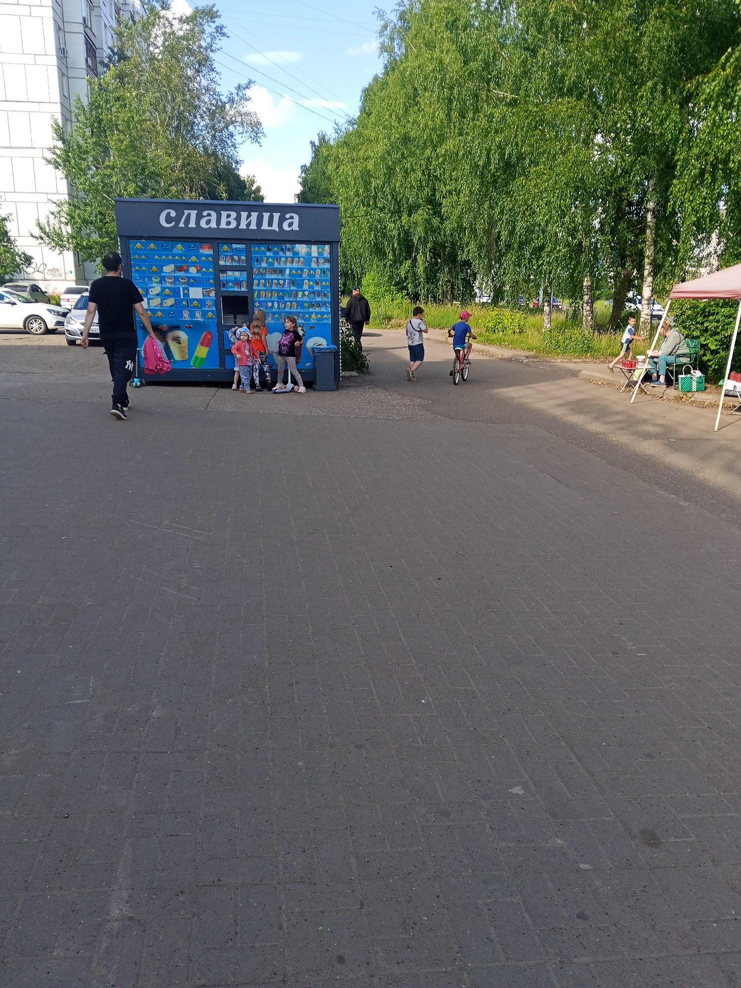 The image depicts an outdoor scene with a street kiosk selling ice cream. The kiosk has "славица" (Slavitsa) written on it. Several people are present, including a man walking away from the kiosk, three young girls standing in front of it, and two boys on bicycles in the background. To the right, a woman is seated under a canopy. The environment appears to be a residential area, with a multi-story building in the background and trees lining the street. The ground is paved with asphalt. The scene suggests a casual, everyday moment in the city of Chaikovsky, Russia, likely during warm weather given the presence of ice cream and outdoor activities.