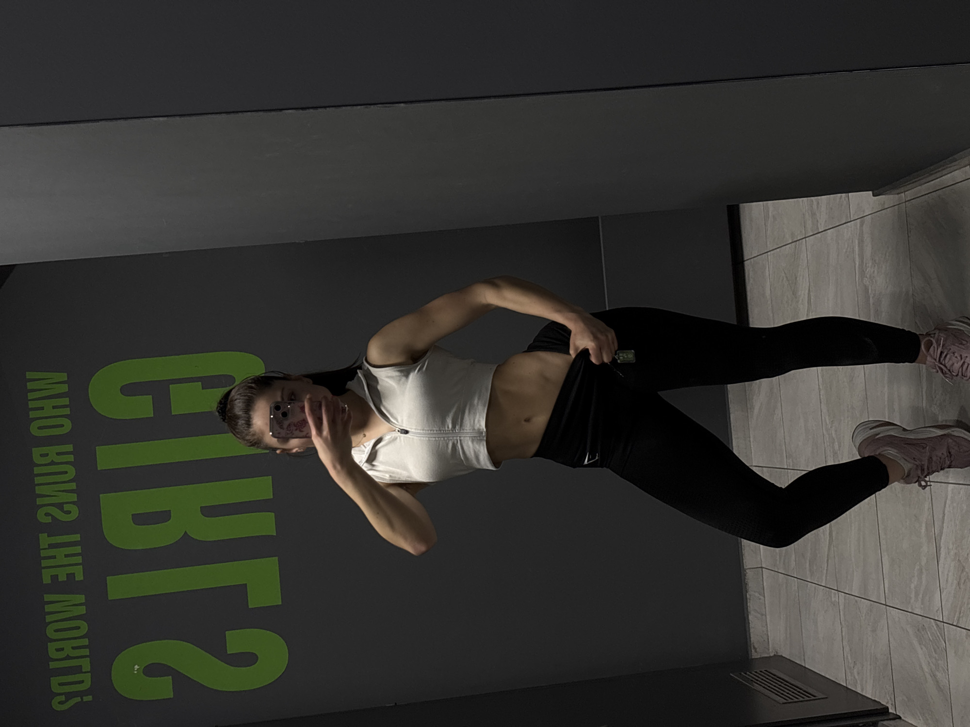 A woman is posing in what appears to be a gym or fitness studio. She is wearing a white zip-up crop top and black leggings, and her pink sneakers are visible in the lower right corner. She is holding her phone up to take a selfie, partially obscuring her face. Behind her, there is a wall with green lettering that reads "GIRLS, WHO RUNS THE MOTORS?". The floor is tiled in a gray pattern, and there's a dark object in the lower right corner that may be part of the gym equipment. The city and specific location are unknown.