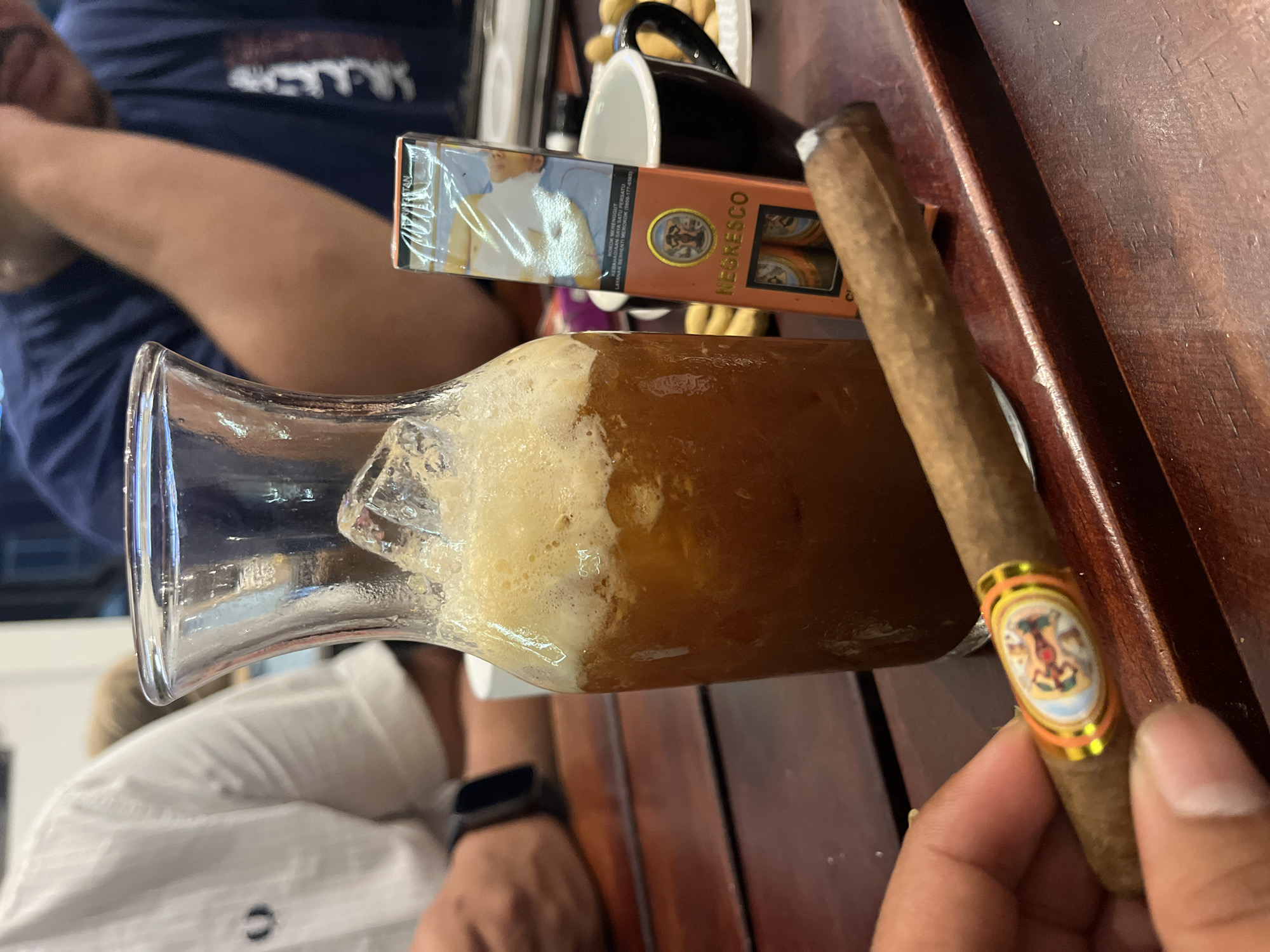The image presents a still life composition on a polished wooden surface, likely a table or bar. Dominating the foreground is a clear glass carafe or pitcher filled with a frothy, amber-colored liquid, possibly beer or a mixed drink. To the right, a lit cigar with a colorful band is held in a person's hand.
Adjacent to the pitcher is a pack of "NEGRESCO" cigarettes. Behind the cigarettes, a black mug with visible contents, possibly snacks, is positioned.
The background includes portions of two individuals: one wearing a white shirt with a dark detail (possibly a logo or button), and another wearing a blue shirt. A wrist with a dark smartwatch is visible.
Although the image doesn't directly reveal the location as Lhokseumawe, Indonesia, the "NEGRESCO" cigarette brand and the presence of individuals suggest an Indonesian setting. Without more context, determining the exact location within Lhokseumawe is not possible.