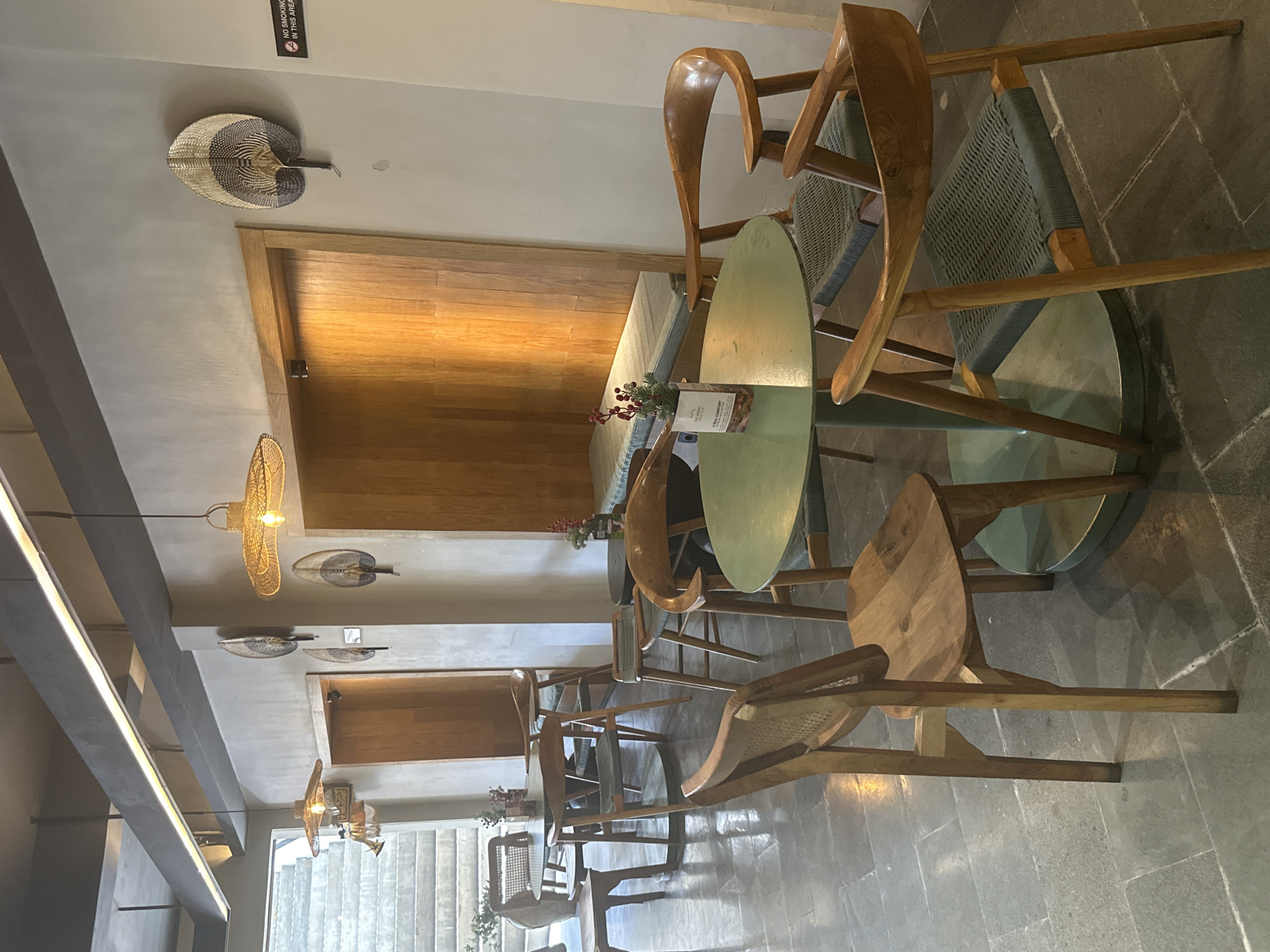 The photograph depicts the interior of a cafe or restaurant in Ubud District, Indonesia. The space is furnished with wooden tables and chairs, set against a backdrop of textured gray walls. Several decorative items are visible, including woven fan-shaped ornaments on the wall and rattan pendant lamps hanging from the ceiling. One notable feature is a built-in wooden bench with cushions, set within a recessed area. A small potted plant with red berries sits on a green, circular table in the foreground, alongside a small informational card. The floor is tiled with large, dark gray stones. The overall ambiance suggests a relaxed and modern aesthetic, with natural materials and warm lighting. There are no people present in the image.