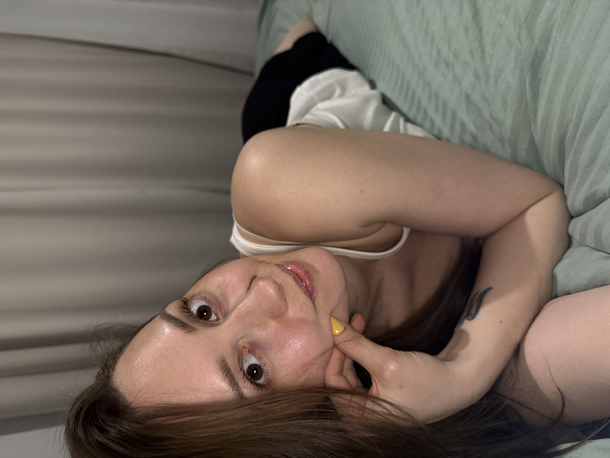 The image depicts a single female subject indoors, lying prone on a bed, facing the camera. She has long brown hair, brown eyes with a wide gaze, and light skin, showing a slight smile. Her right hand, adorned with yellow nail polish on the visible fingernails, supports her chin/cheek. A dark-colored tattoo, possibly a wing or feather design, is visible on her right forearm. She is wearing a white tank top, and a black garment is visible on her lower body. The bed is covered with light green sheets featuring a subtle striped pattern. The background consists of light brown/beige curtains or drapes. The specific location in the city of Chaikovsky, Russia, cannot be confirmed from the visual evidence within the image.