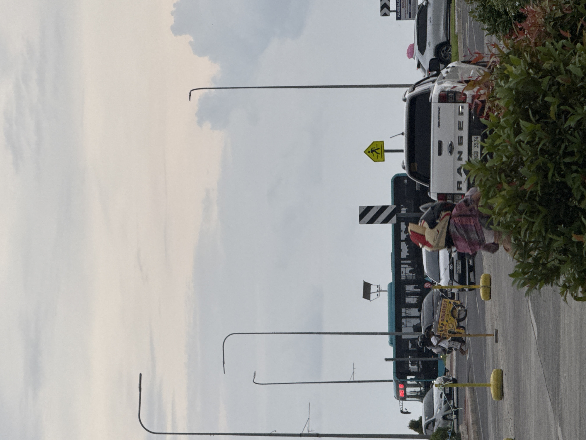 The image captures a daytime street scene in Davao City, Philippines, under an overcast sky. The perspective looks down a multi-lane road, showing a mix of vehicles and urban infrastructure.
In the mid-ground, a white Ford Ranger Wildtrak pickup truck is prominently featured, its rear facing the viewer. The truck's license plate, "NGO 3534," is clearly visible. Partially obscured by a large green bush with reddish leaves in the right foreground, a person is visible near the pickup truck, wearing a plaid-patterned garment and a light-colored top or hat. Behind the pickup, another white SUV is parked.
Further down the road, a long, blue and green bus occupies the left lane, its front digital display showing unreadable red numbers. Several other cars are visible along the street. On the right side of the road, a vendor is pushing a yellow cart that displays the partially visible word "GREATEST." Yellow bollards or markers delineate the roadside.
Numerous road signs dot the scene, including a bright yellow triangular sign with a black pedestrian icon, indicating a pedestrian crossing. Another black and white chevron sign has visible text, part of which reads "PAMBALO." Tall streetlights arc over the road, extending towards the sky. The light is diffused and soft, suggesting an overcast day rather than direct sunlight, with no harsh shadows visible.