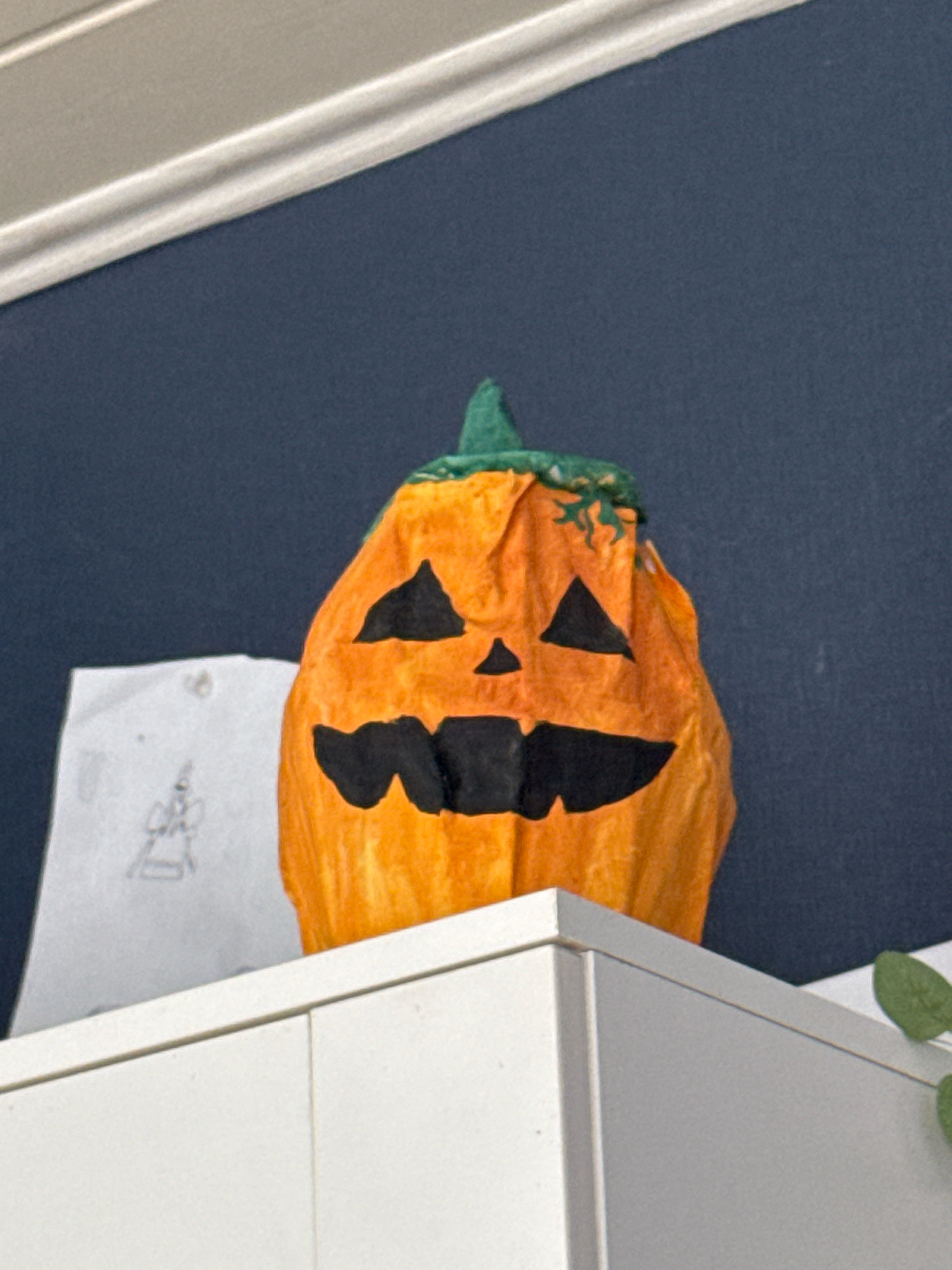 The image shows a handmade Halloween pumpkin decoration. The pumpkin is orange with a green stem and has a carved face with triangular eyes and a jagged mouth, all colored black. It appears to be made from paper or fabric, with visible wrinkles and creases. To the left of the pumpkin, a piece of white paper with a simple drawing of a figure is partially visible. The pumpkin is placed on top of a white surface, possibly a shelf or cabinet, with a dark blue wall in the background. A white molding or trim runs along the top of the wall. A small green leaf from a plant is visible in the bottom right corner of the image. The image is taken from a slightly upward angle. The scene is indoors. The image does not provide any information that would allow for the determination of its location in Überherrn, Germany.