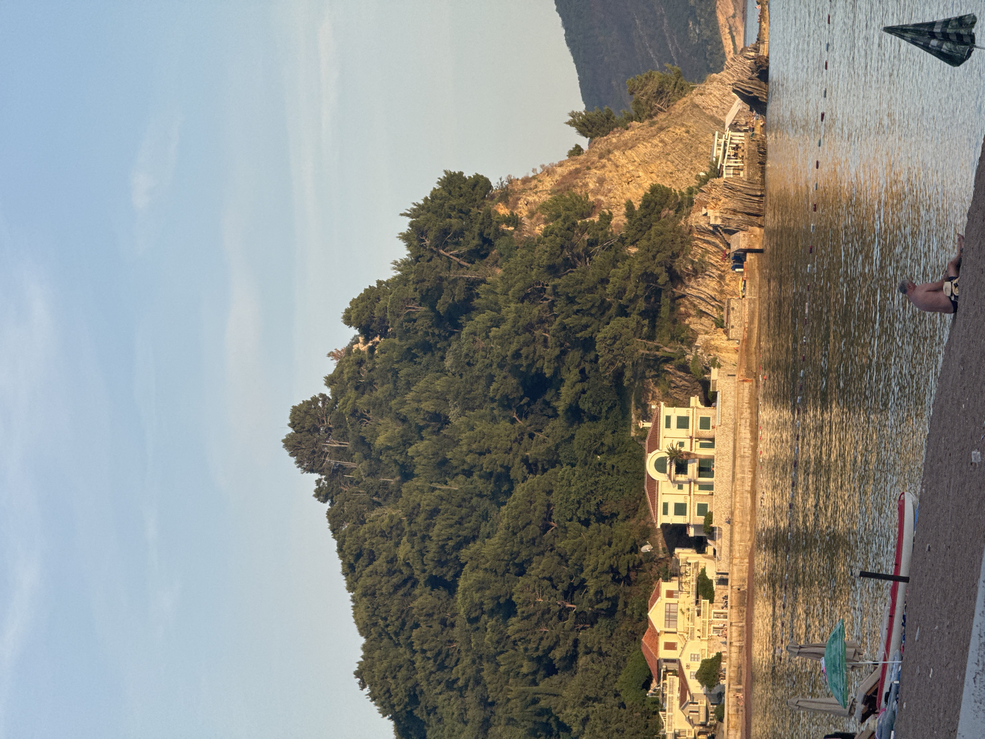 The image shows a coastal scene in Petrovac centar, Montenegro. A large, tree-covered hill slopes down to the shoreline, where several buildings are situated. The architecture of the buildings suggests residential or resort structures. The water's surface is reflective, indicating calm conditions. A checkered flag stands at the edge of the water, near a concrete barrier, where a person is sitting. In the foreground, there are a few beach chairs and umbrellas.