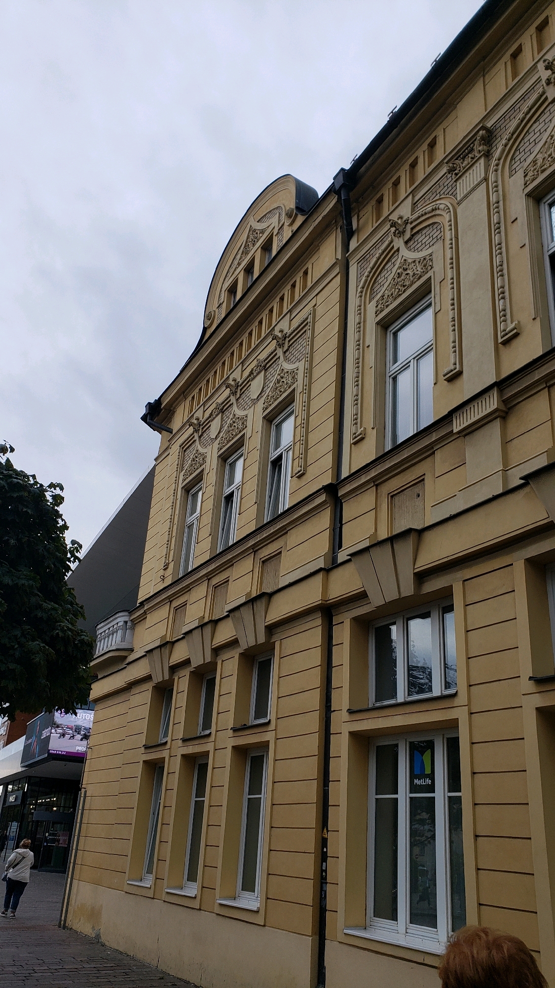 The image shows a section of a light yellow, ornate building on a street in Prešov, Slovakia. The building features classical architectural details, including decorative moldings around the windows and along the roofline. Several tall, rectangular windows with white frames are visible, some of which are open. One window on the lower floor displays a MetLife logo. Adjacent to the yellow building on the left, a modern, dark grey structure with a large advertisement billboard is partially visible. A tree with dark green foliage is also present on the left side of the image. A woman in a light-colored jacket and dark pants is walking on the paved sidewalk in front of the buildings. The sky is overcast and grey.