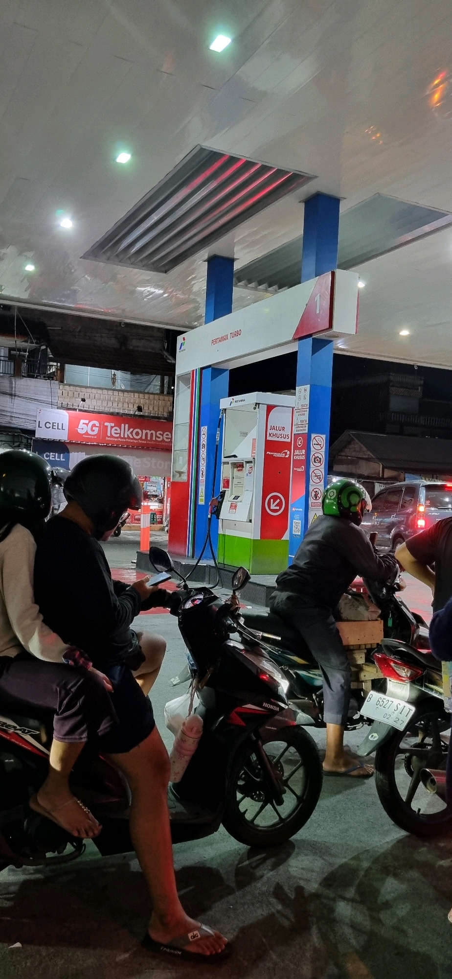 At a Pertamina gas station in Denpasar, Indonesia, several individuals on motorcycles are visible under the station's canopy. The scene is lit by overhead lights and the station's illuminated signage. In the foreground, two people on a scooter are facing away from the camera. The rider, wearing a black helmet, is looking at their phone. Behind them, another person, also wearing a black helmet, is seated on the scooter.
Further into the scene, a person wearing a green helmet is seated on another motorcycle, facing towards a gas pump. Their right hand is near the gas pump. A third motorcycle, with its license plate partially visible as "B 6527 SIT", is also present. In the background, other vehicles and buildings can be seen. The gas station features prominent red and white branding, with "PERTAMAX TURBO" displayed on a sign. A sign reading "JALUR KHUSUS" (Special Lane) is also visible on the gas pump. The overall atmosphere suggests a typical evening at a busy urban gas station.