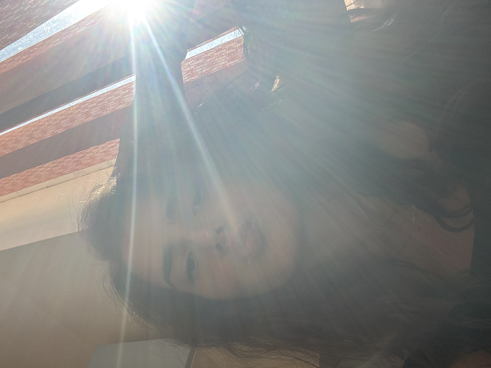 A young woman with long, dark hair is captured in a selfie, her face partially obscured by bright sunlight streaming through vertical blinds. She is making a kissing gesture, with her lips puckered. The lighting is strong, creating lens flares and blowing out some details of the image. The background suggests an indoor setting with a window covered by blinds, possibly in a residential area of Shumen, Bulgaria.
