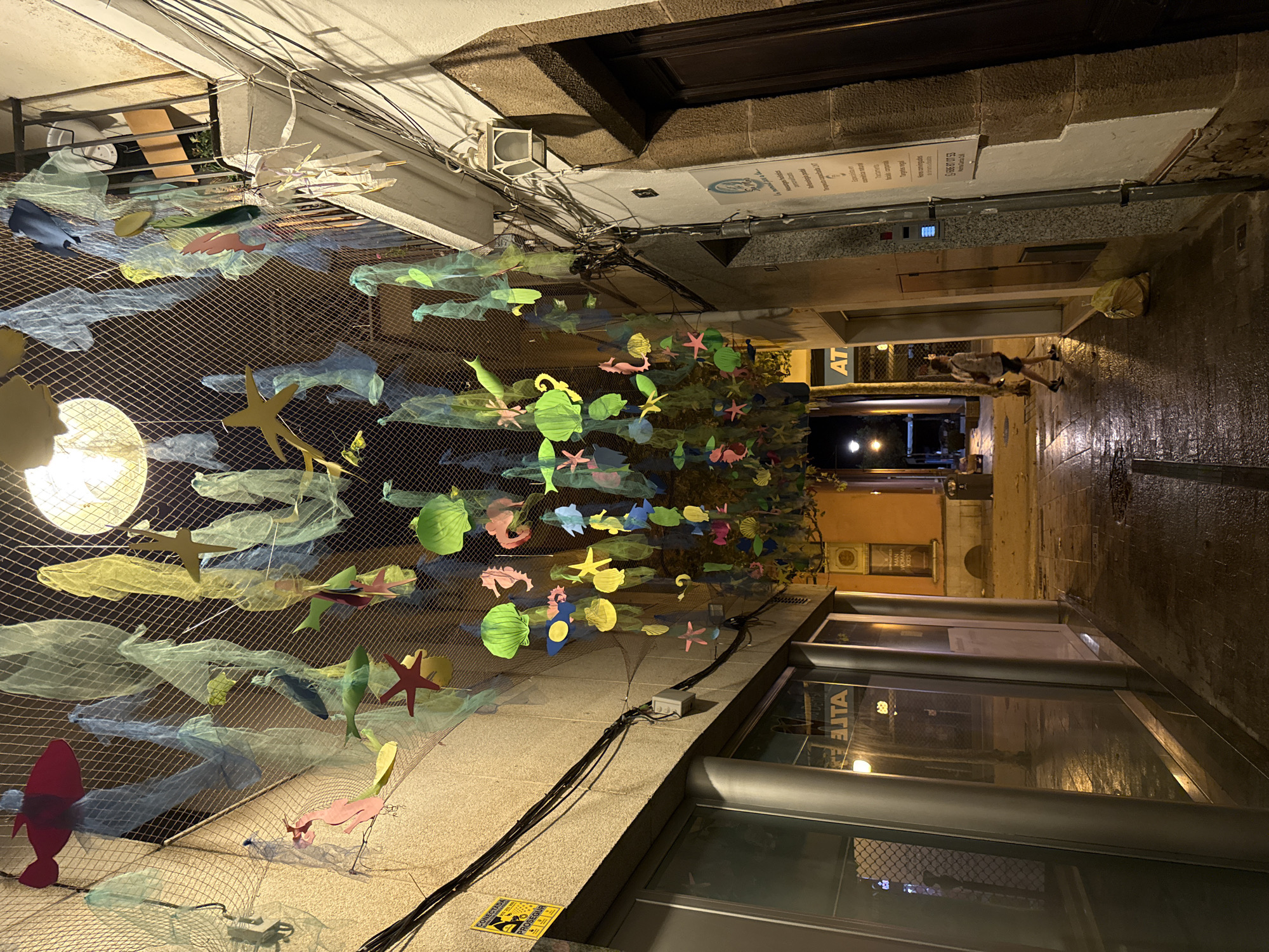 This image captures a narrow street at night in Blanes, Spain, adorned with a decorative overhead installation of fabric and paper cutouts resembling sea life, such as fish, starfish, seahorses, and shells. The colorful sea creatures are suspended from a net, creating a whimsical, underwater effect. The street itself is paved and appears wet, suggesting recent rain, with reflections visible on the ground. On the left, a building with large glass windows, possibly a storefront, is illuminated from within. A sign with Spanish text is partially visible. On the right, a building with a textured facade features a wooden door and a mounted lantern. Electrical wires and conduits are visible along the building's exterior. Further down the street, a single person, a woman with light-colored clothing and a backpack, walks away from the camera. A yellow trash bag lies on the ground near her. The overall ambiance is that of a quiet, atmospheric evening in a European town.